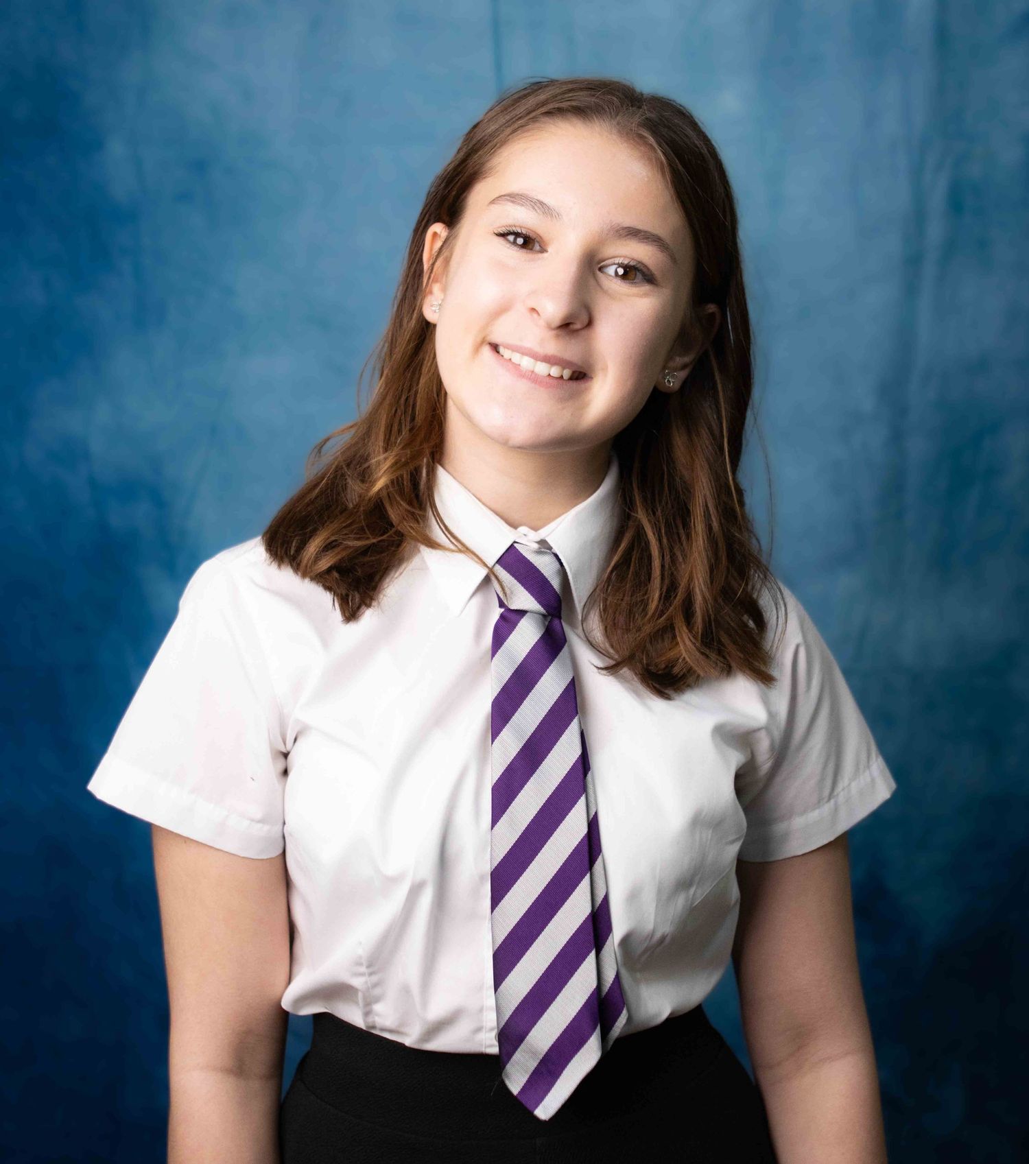 Student in school uniform with white shirt and purple striped tie poses for portrait against blue backdrop.