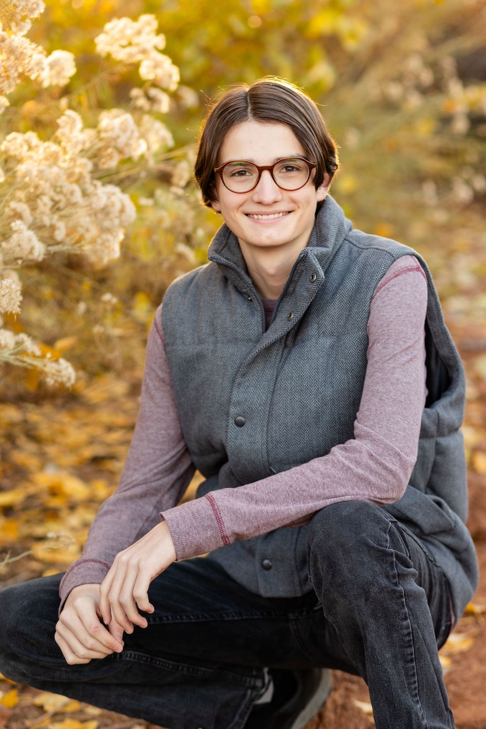young man with round glasses and puffy vest smiles at the camera while in a crouched position