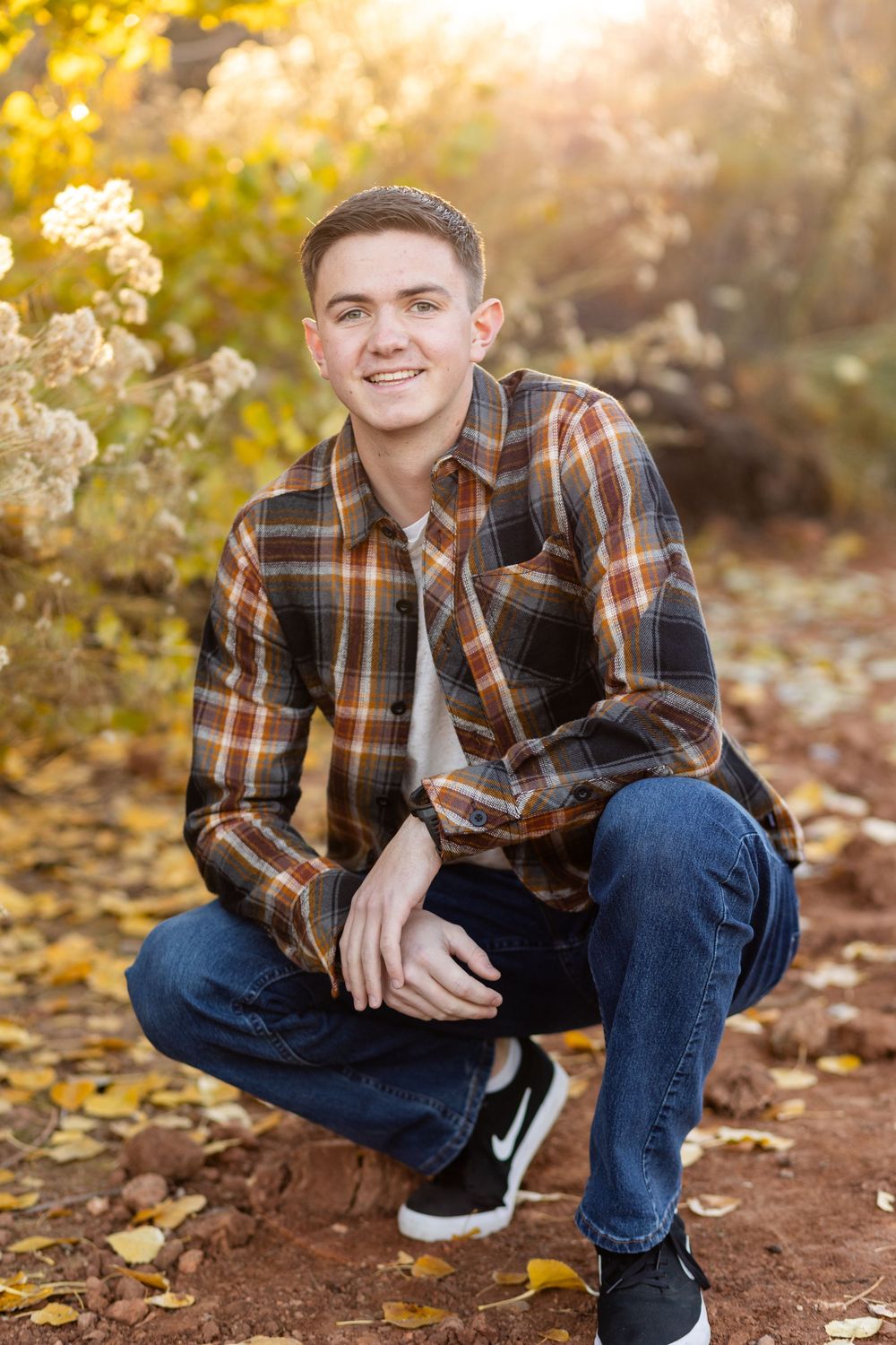 young man in a crouched position wearing a plaid shirt smiling at the camera with faded yellow blooms