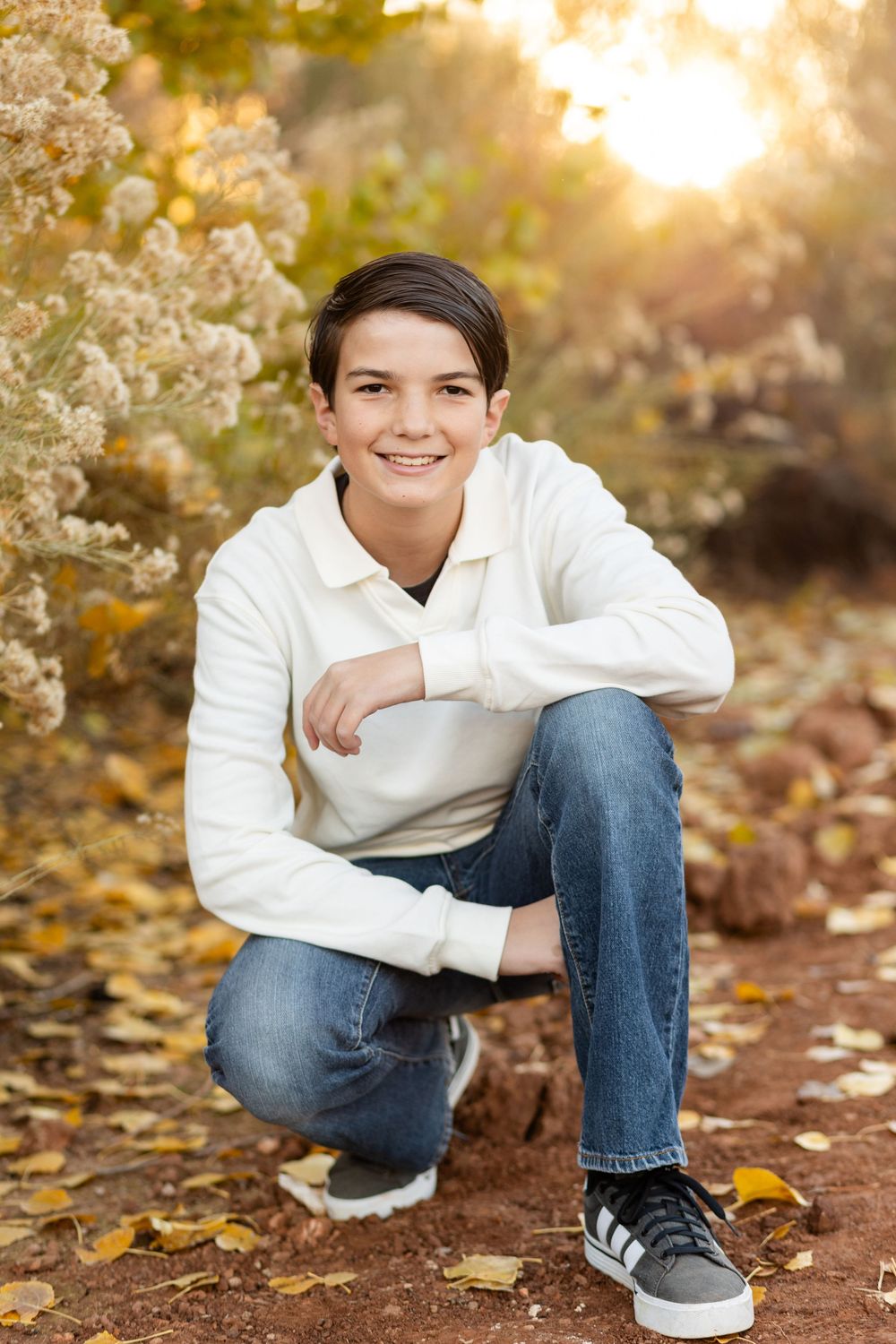 young man in a white sweater crouched smiling at the camera with faded yellow blooms