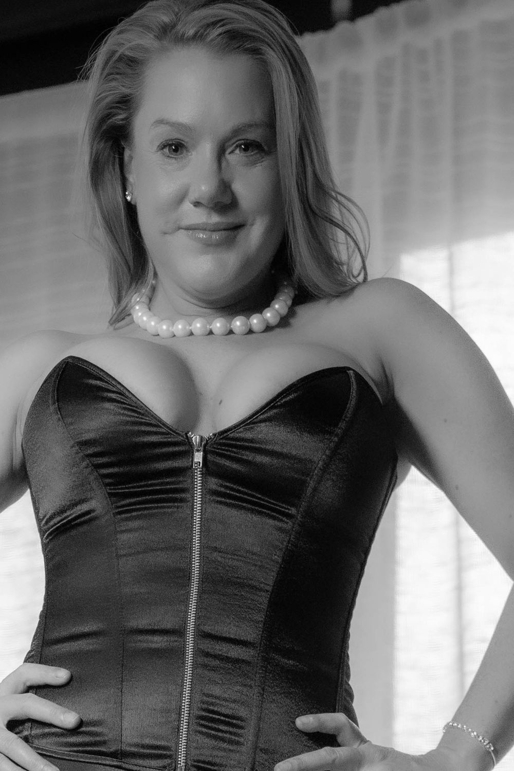 Person wearing black corset top with pearl necklace in front of white curtains.