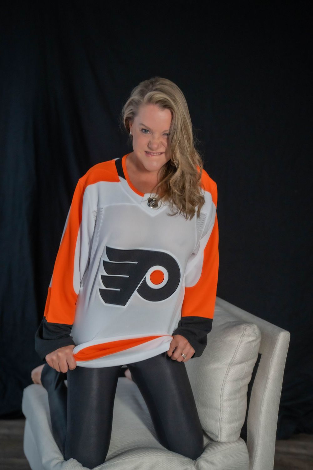 Person wearing Philadelphia Flyers hockey jersey posing on white chair against dark background.