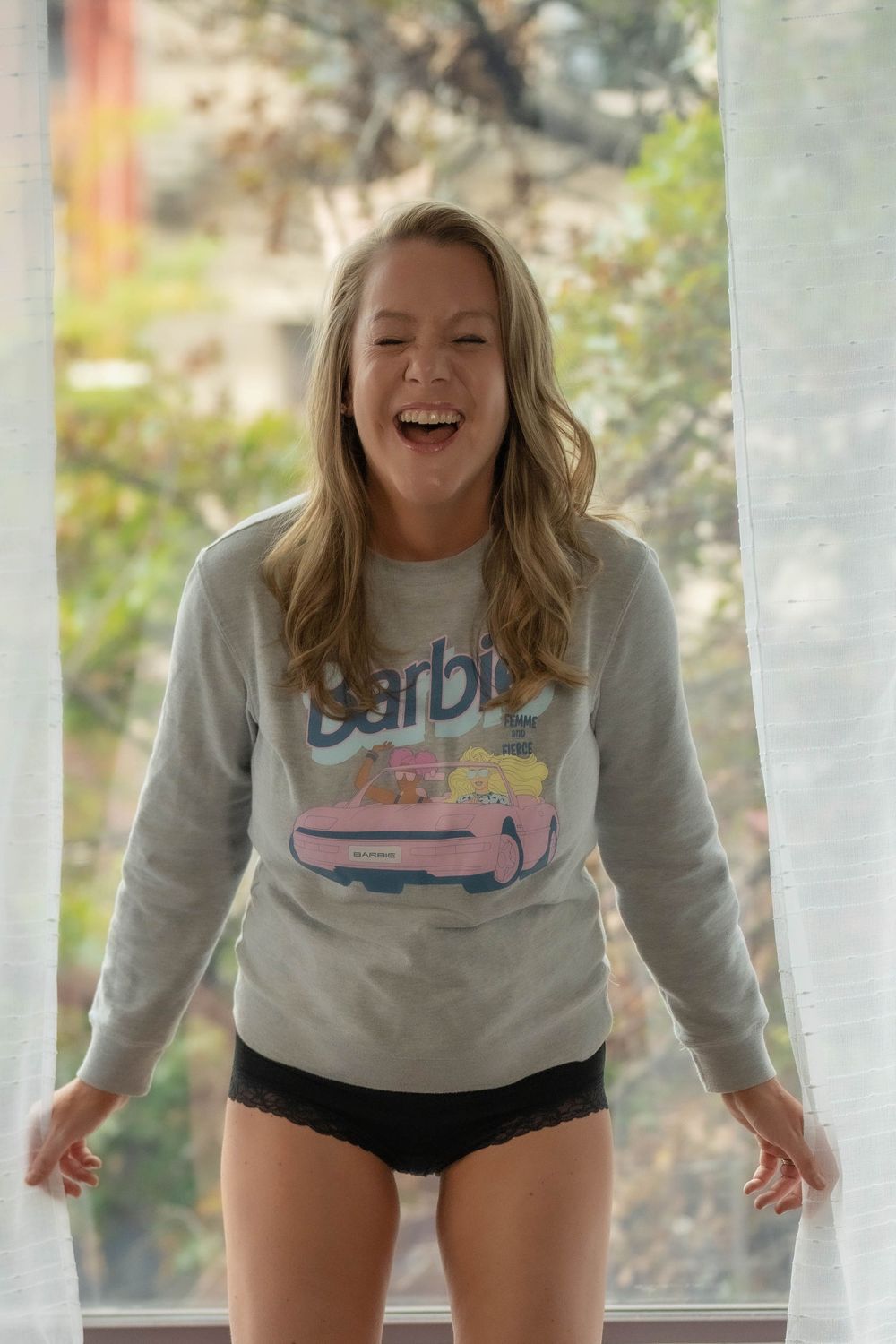 Person in grey sweatshirt and black shorts laughing and posing on a porch with autumn foliage in background.