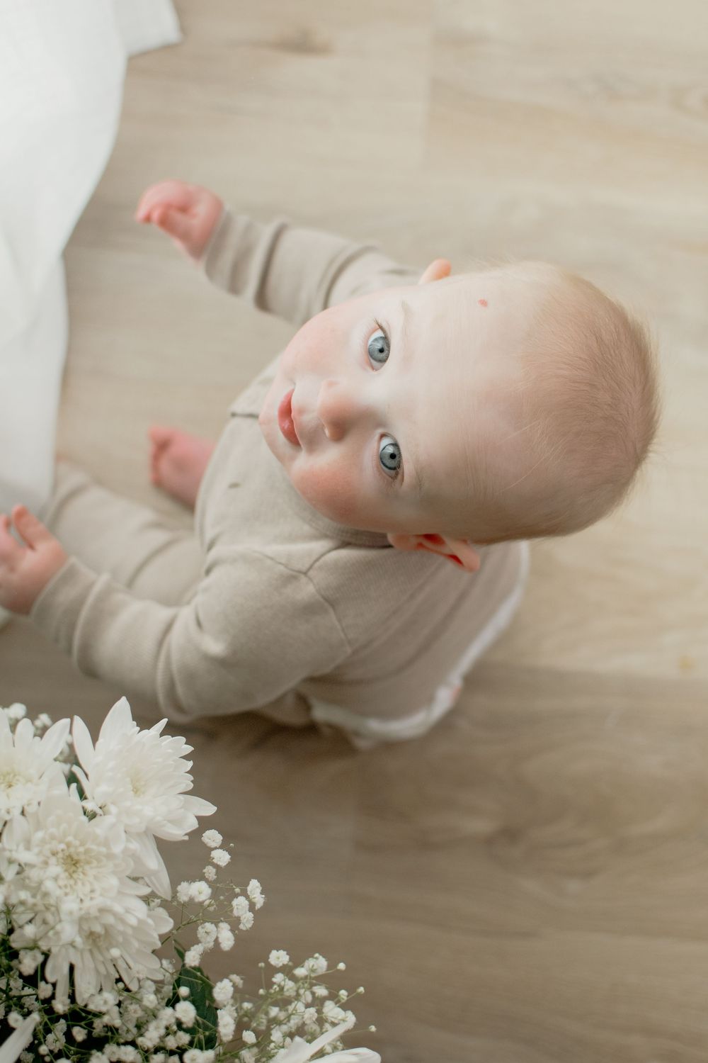 A baby in light clothing lies on wooden flooring next to white flowers and baby's breath floral accents.