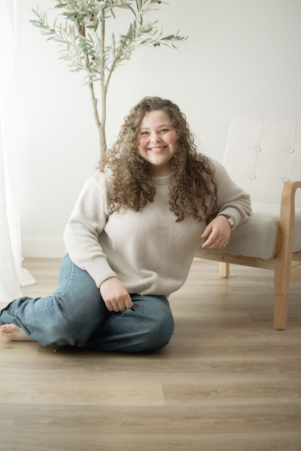 A person in a cream sweater and jeans sits relaxed on a wooden floor with neutral decor and plant accents.