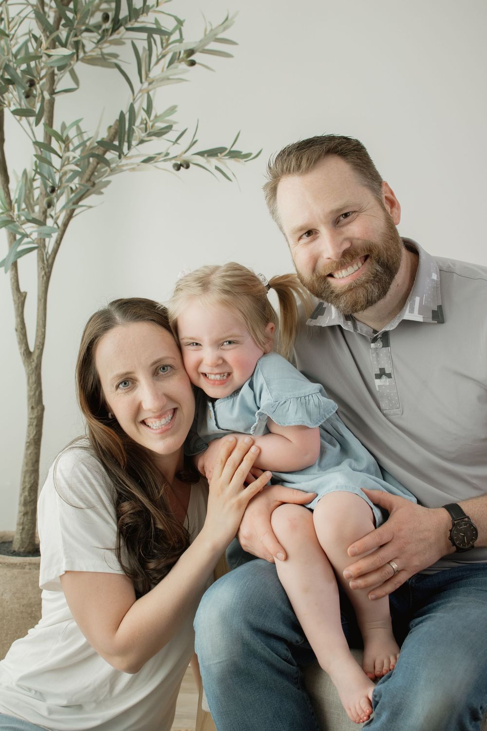 A family with a young child poses for a series of candid photos against a light-colored wall with plant decor.