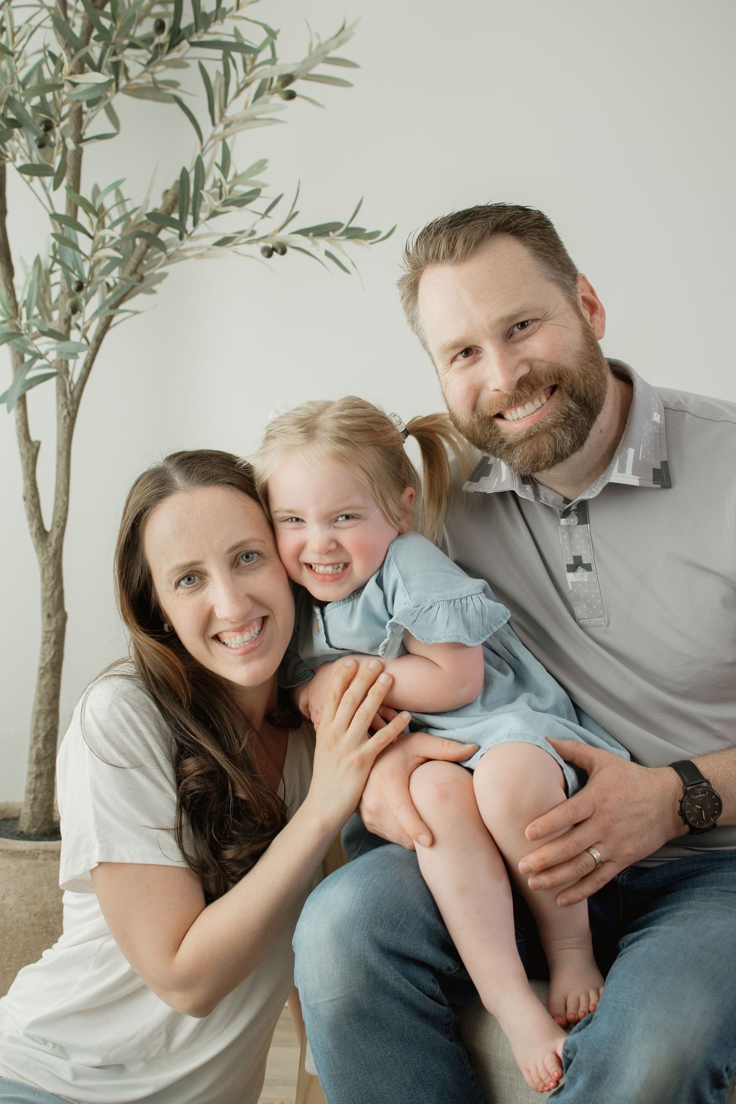 A family with a young child poses for a series of candid photos against a light-colored wall with plant decor.