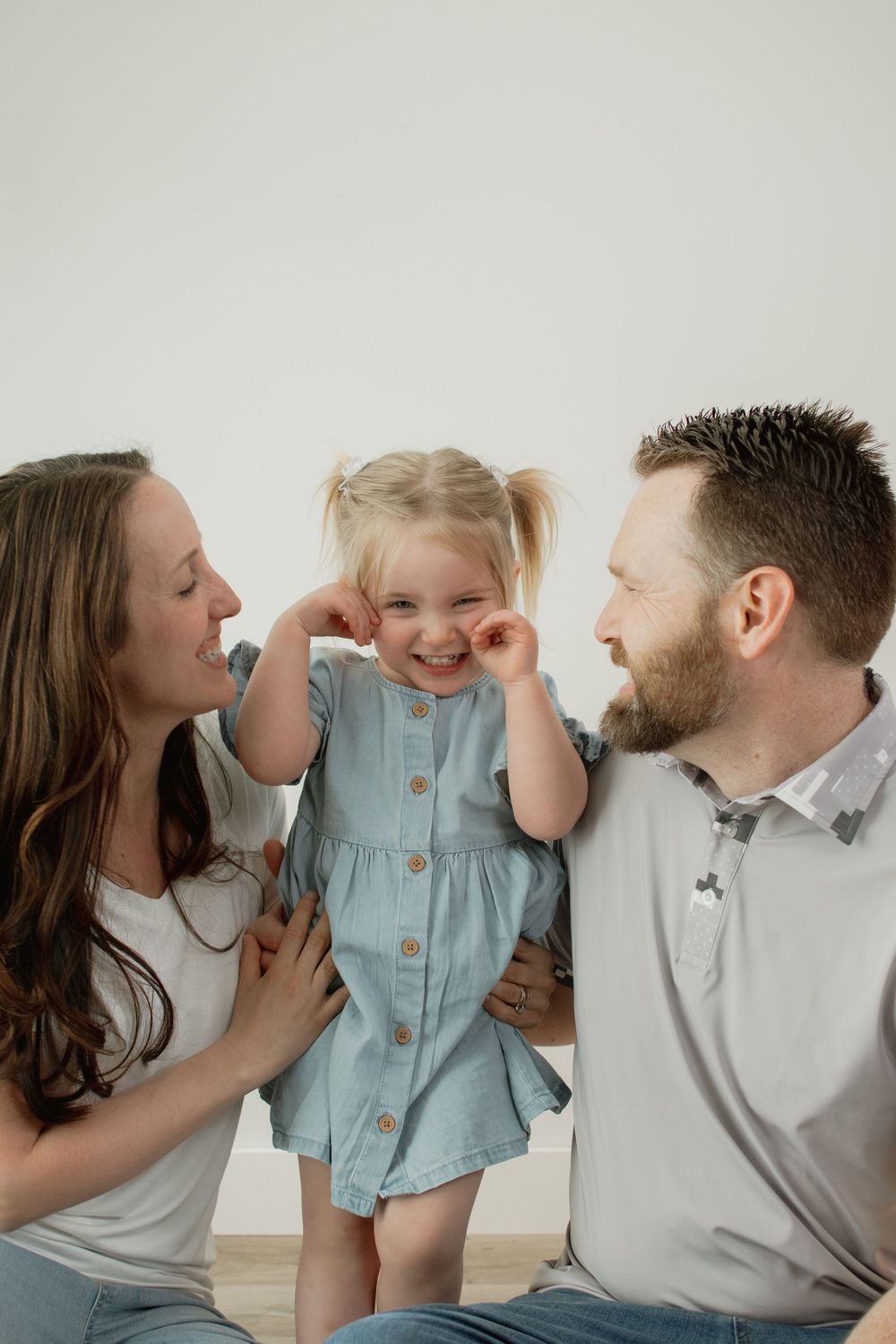 A joyful family moment shows parents kissing their small child on both cheeks against a light background.