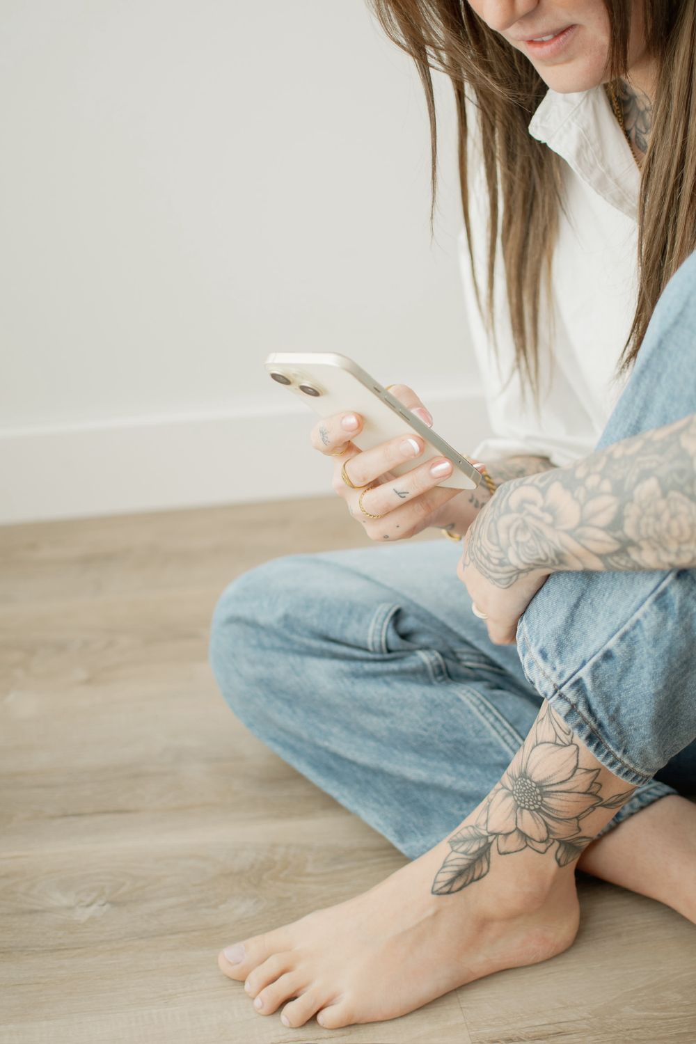 Person in jeans with floral tattoos sits on hardwood floor while checking their smartphone.