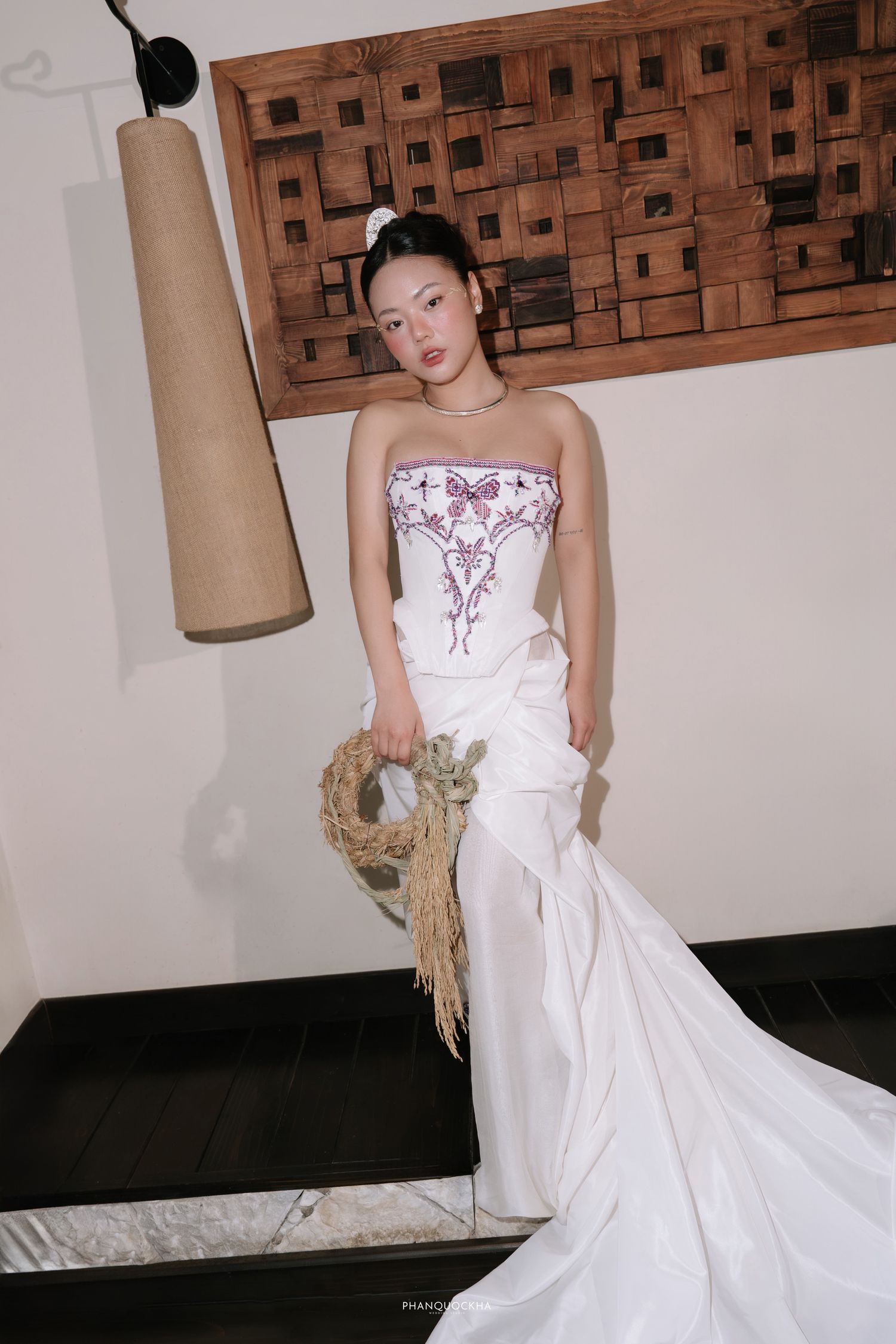 Bride in white strapless gown with floral embroidery holding dried wreath bouquet indoors.