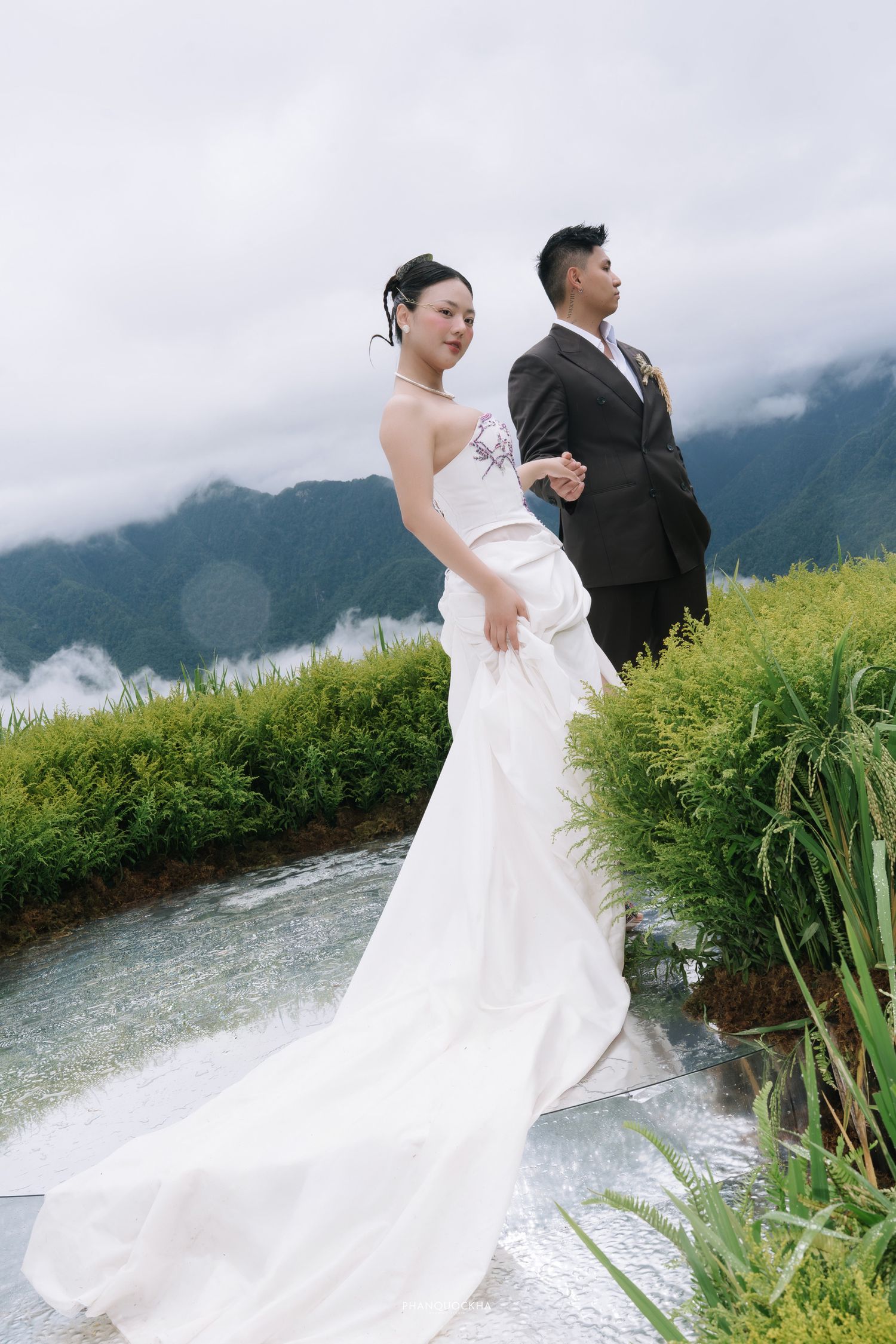 Elegant bride in strapless gown and groom stand hand-in-hand on rice terrace with mountain views.