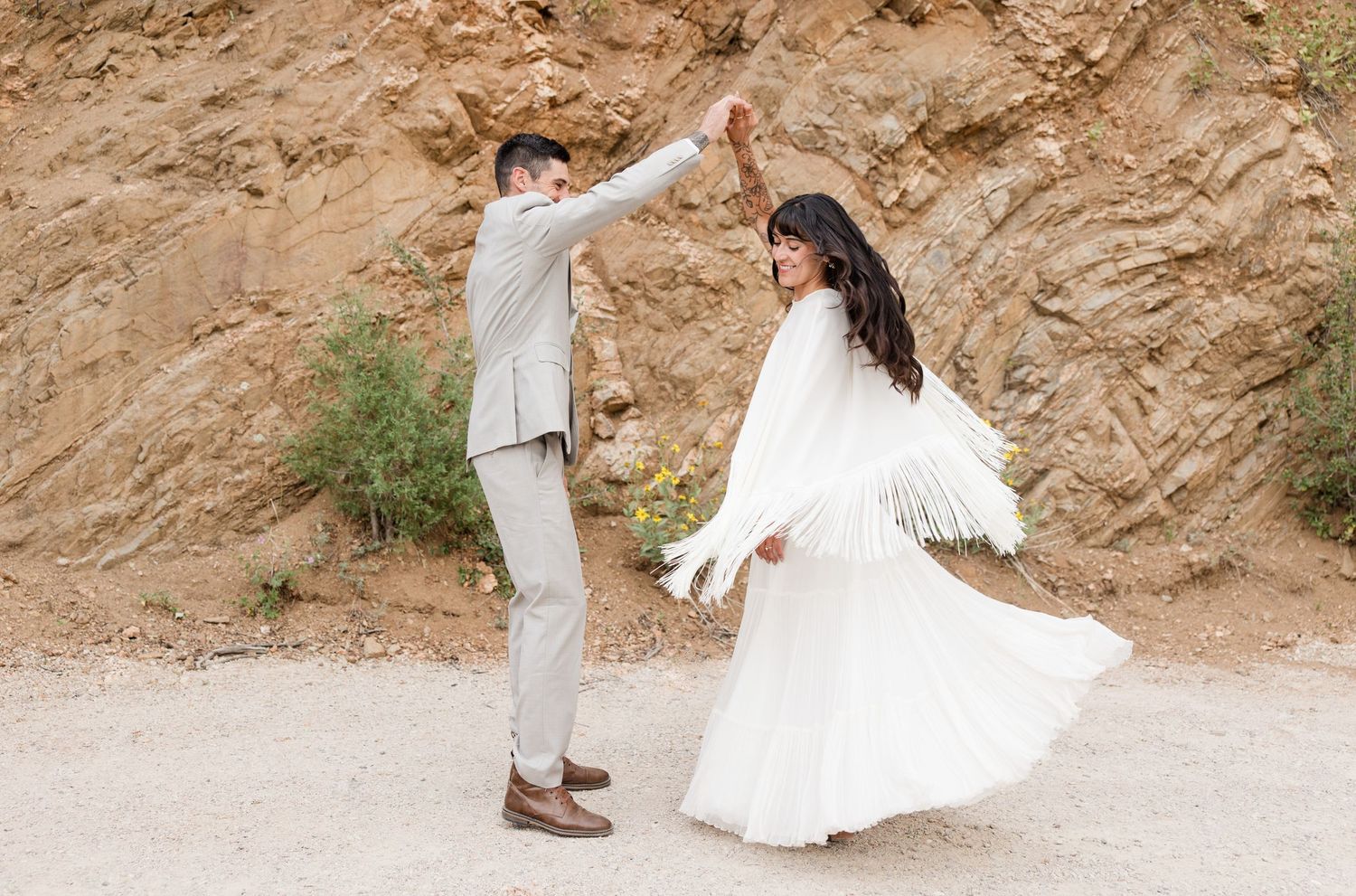 A couple dances together on a dirt path, with the bride wearing a flowy white dress with feathers against an earthy backdrop.