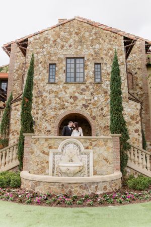 Couple kissing by a stone fountain at a Tuscan-style wedding venue with cypress trees and floral gardens.