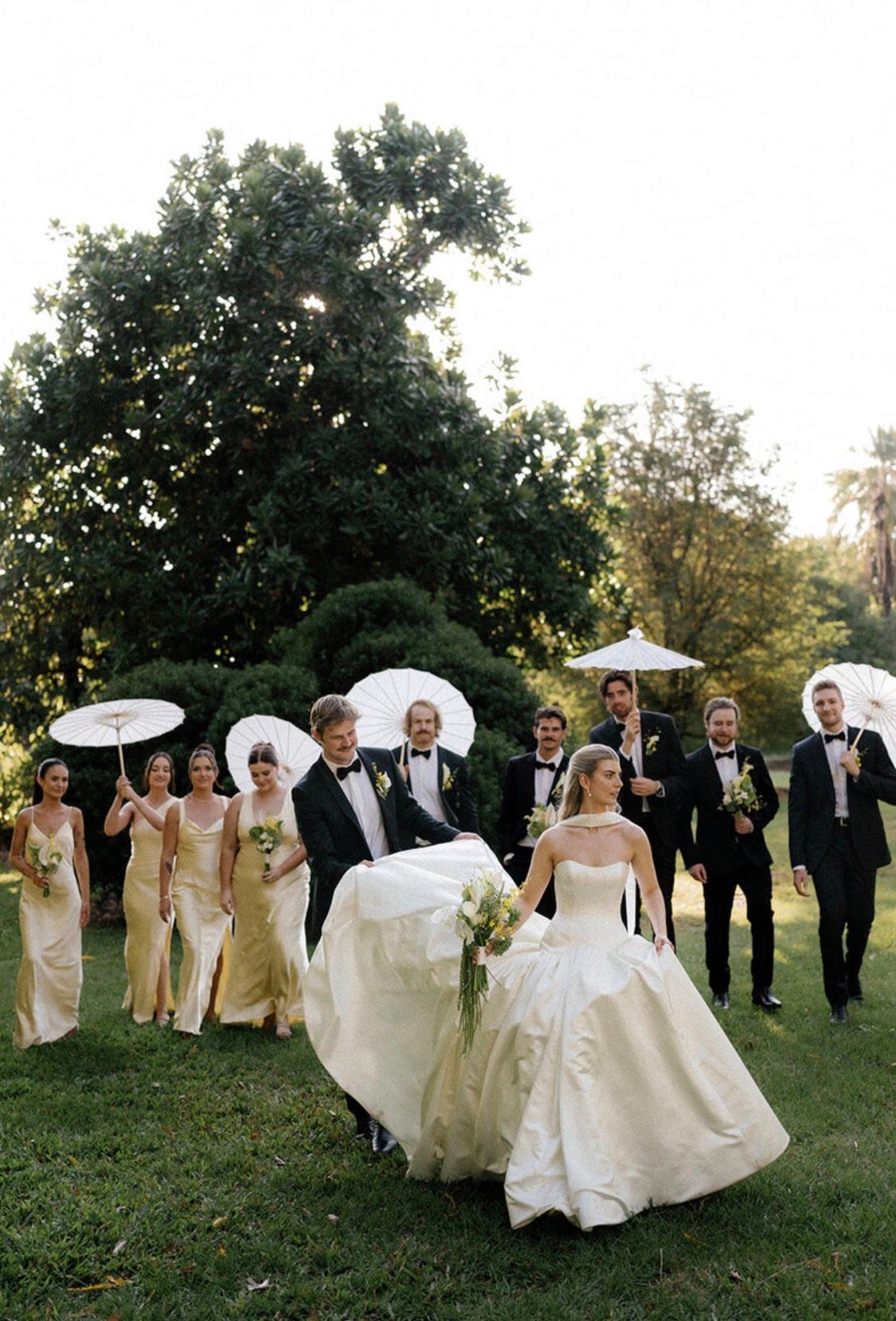 Wedding party on green lawn, bride in white gown carried by groom, bridesmaids in gold dresses holding white parasols, groomsmen in tuxedos.