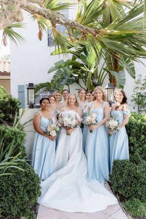 Bridesmaids wearing light blue dresses pose with bride in white gown under tropical palm trees at outdoor garden venue.