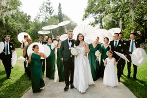 Wedding party in emerald green dresses and black suits pose with decorative white parasols on a garden path.