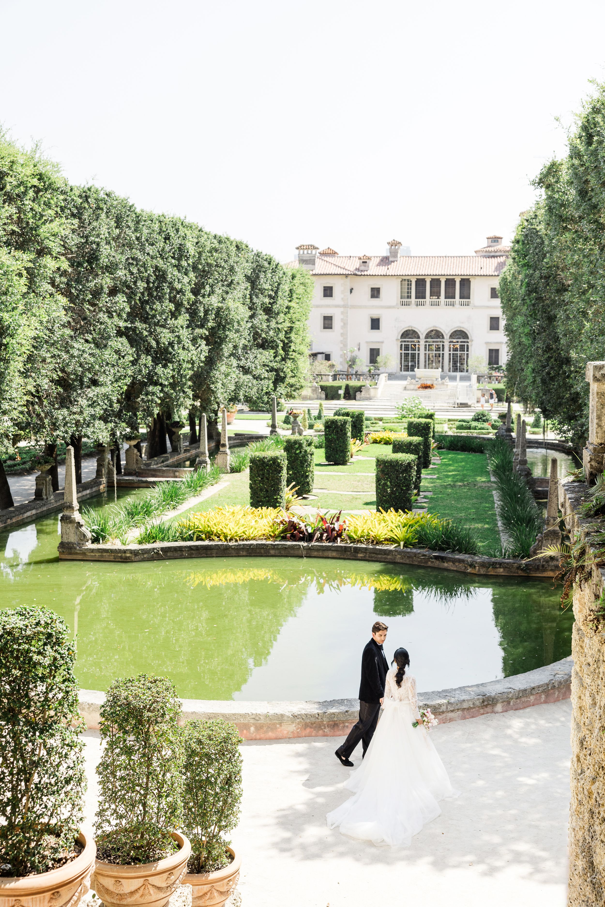 Elegant couple walking through manicured garden with mansion and reflecting pool.