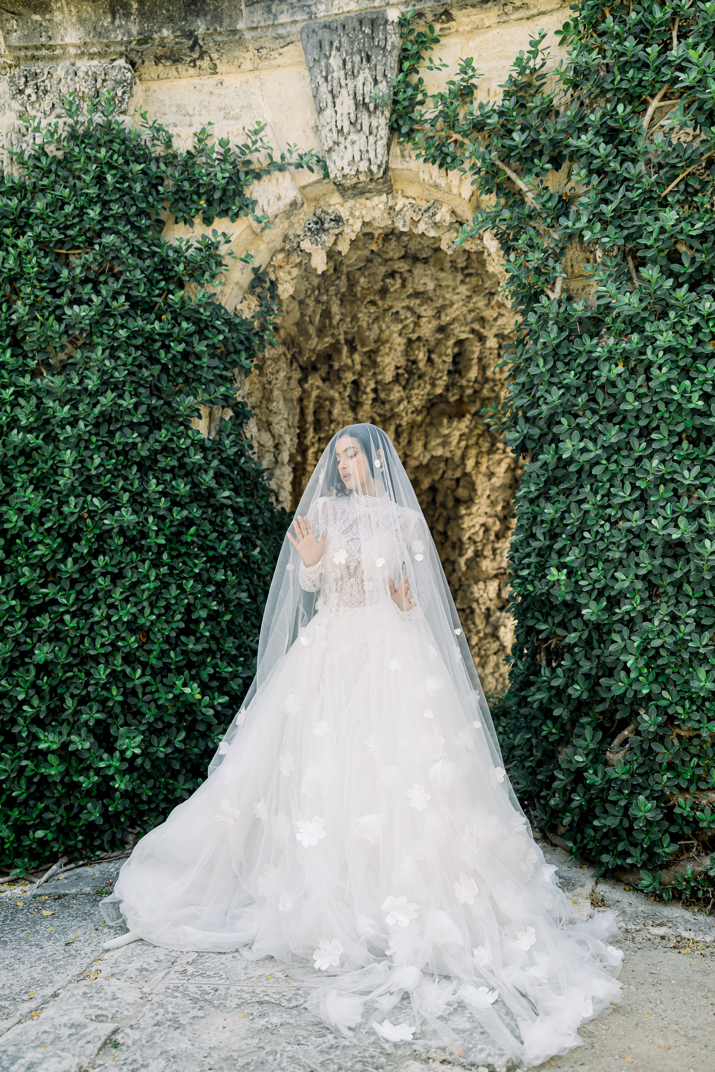 Bride in flowing white gown and veil poses in garden grotto.
