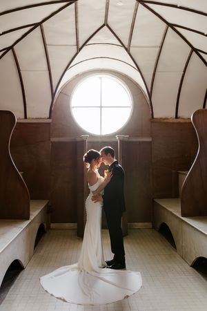 Bride and groom share an intimate moment in a chapel, backlit by a round window with arched ceiling.