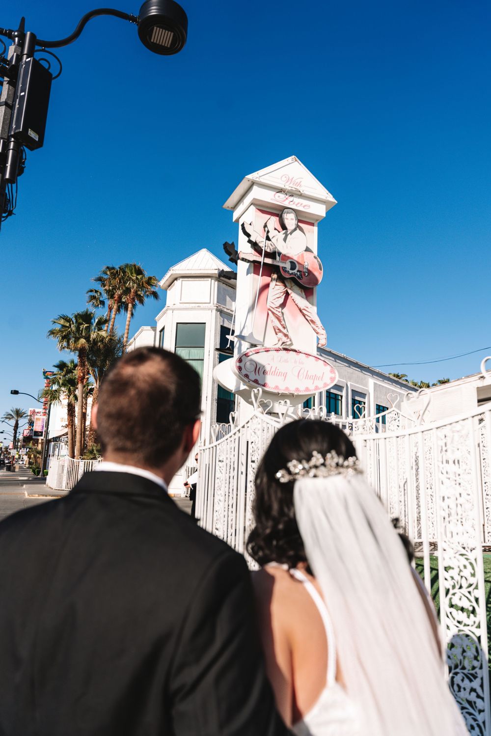 The back of people in wedding attire looking at a white gate entrance with palm trees and blue sky behind.