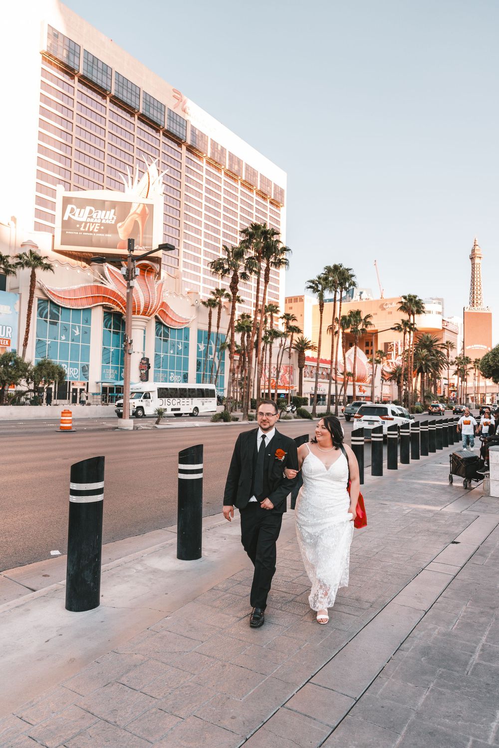 Wedding couple walking down palm-lined sidewalk outside convention center building in warm sunlight.