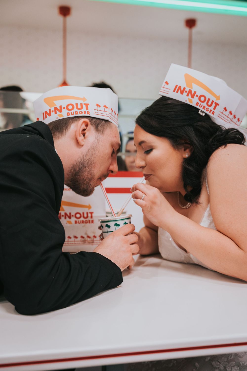Couple in In-N-Out Burger paper hats share a milkshake together across a white table in a romantic moment.