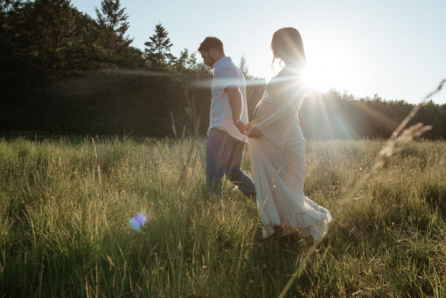 Two people walk through a sunlit meadow of tall grass at golden hour while holding hands.