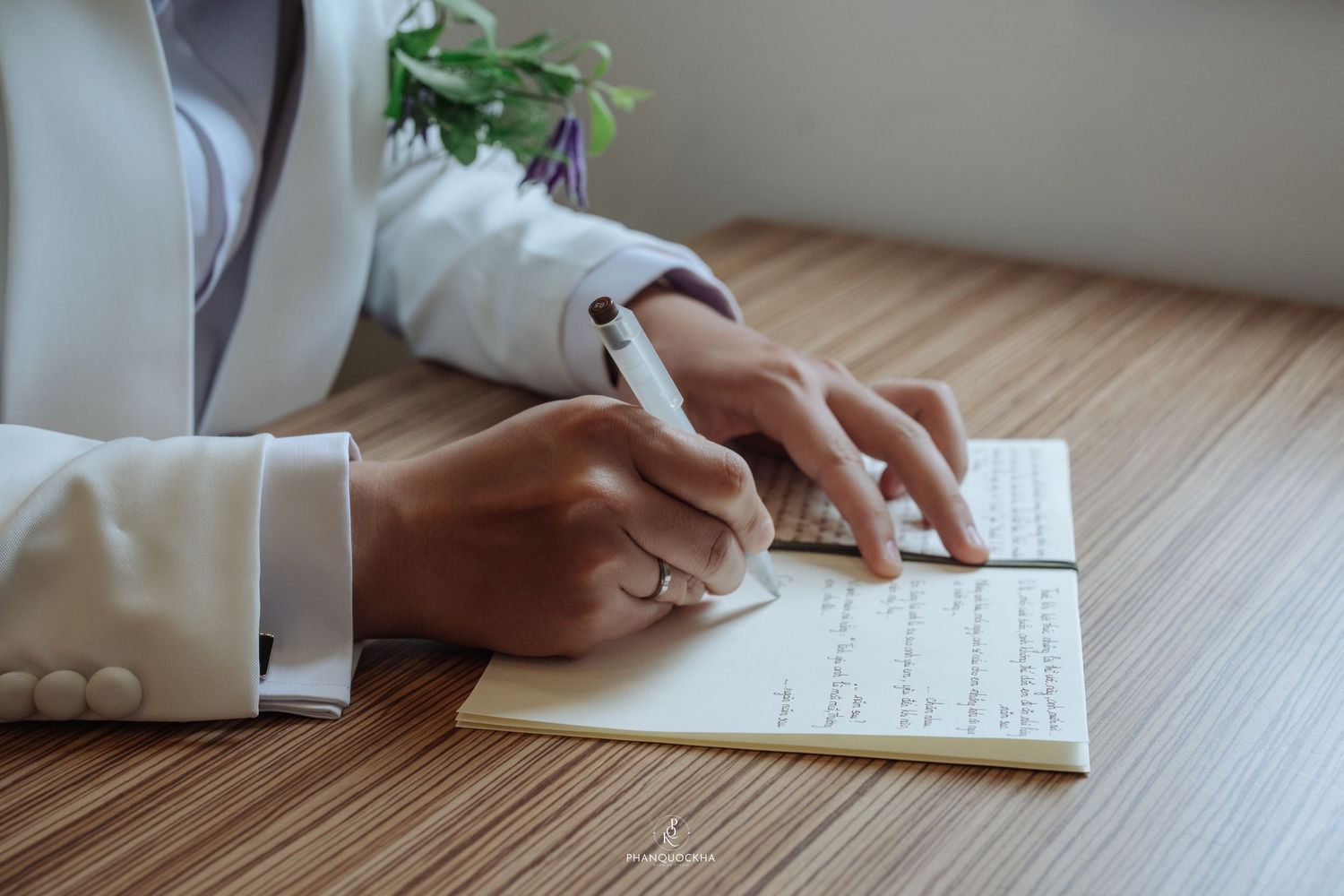 Close-up of person signing wedding registry with pen wearing white suit and ring.