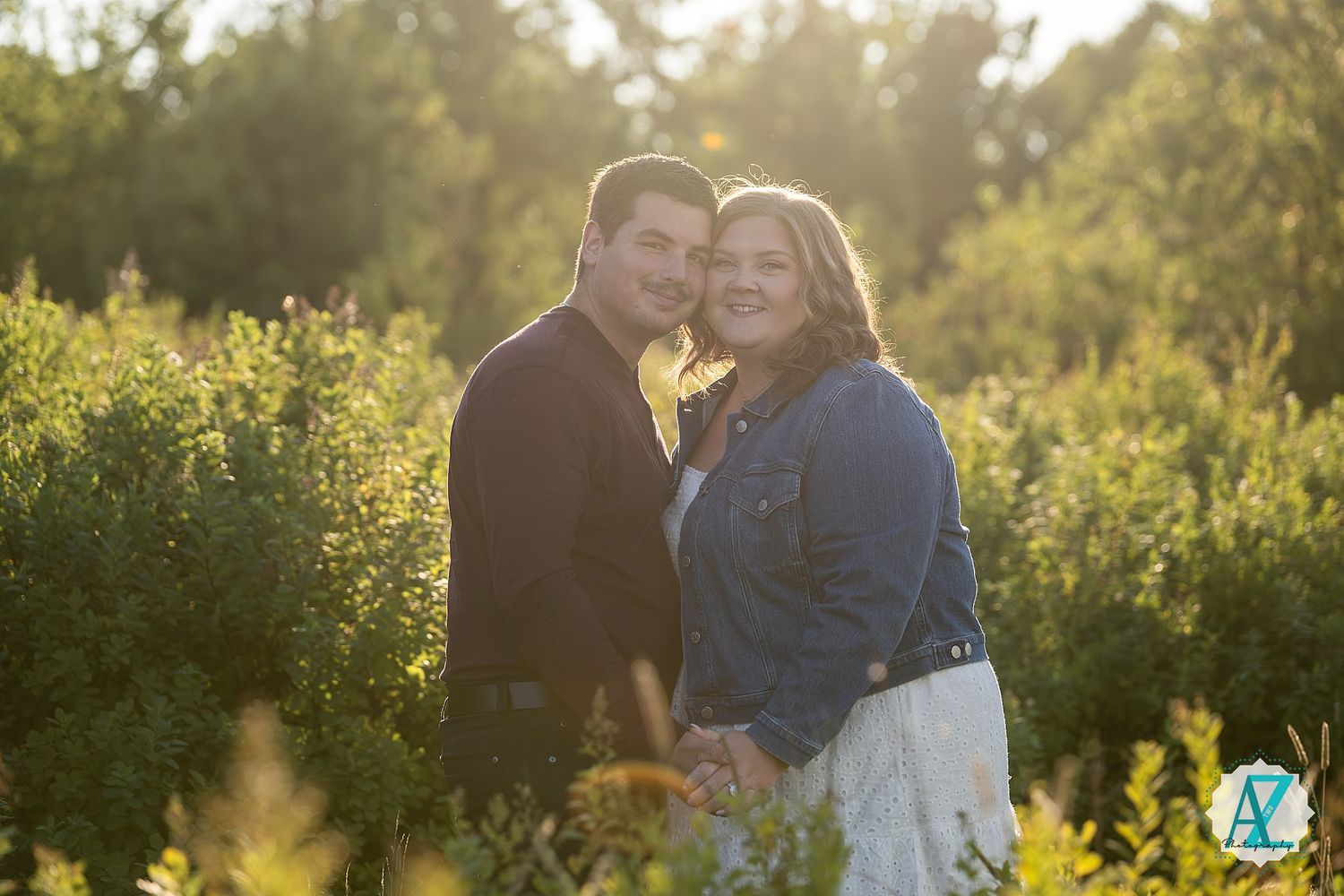 A couple poses together in a sunlit field at golden hour wearing casual attire.