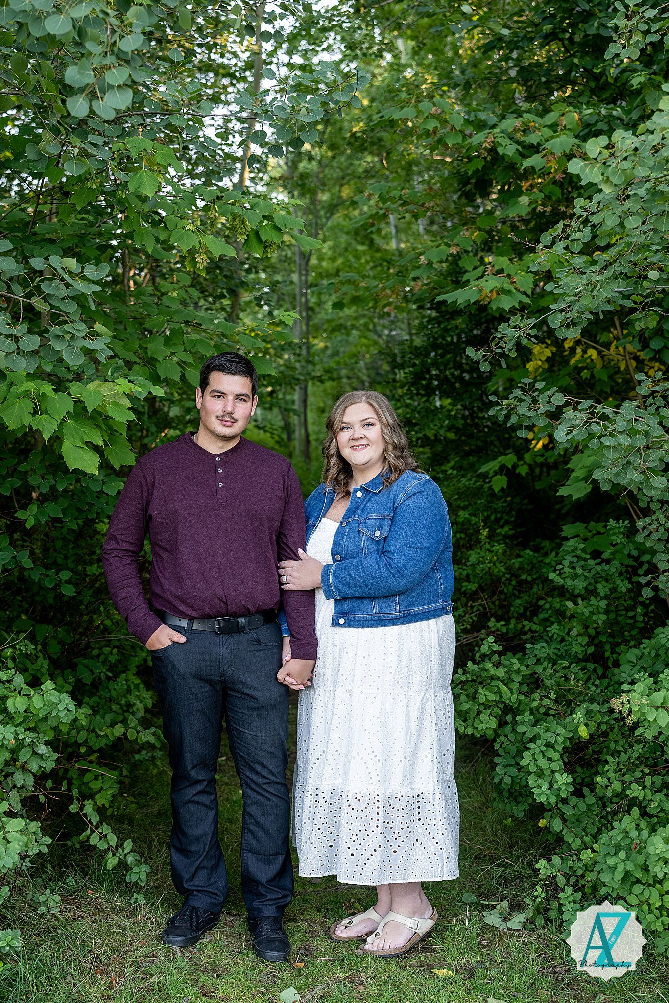 Couple wearing purple and denim outfits pose together against lush green foliage background in outdoor portrait session.