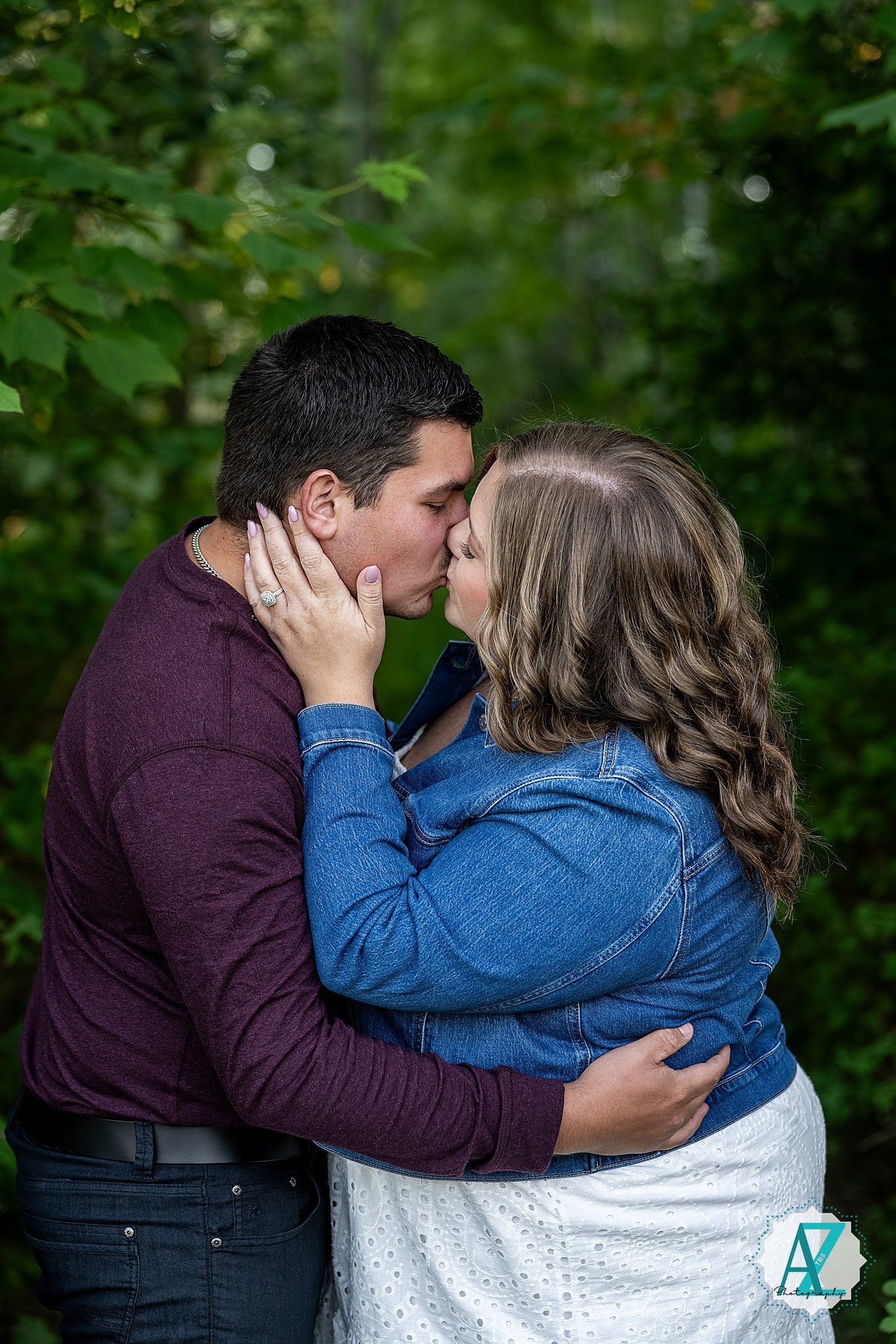 A couple in casual attire sharing a romantic embrace and kiss outdoors among green foliage.