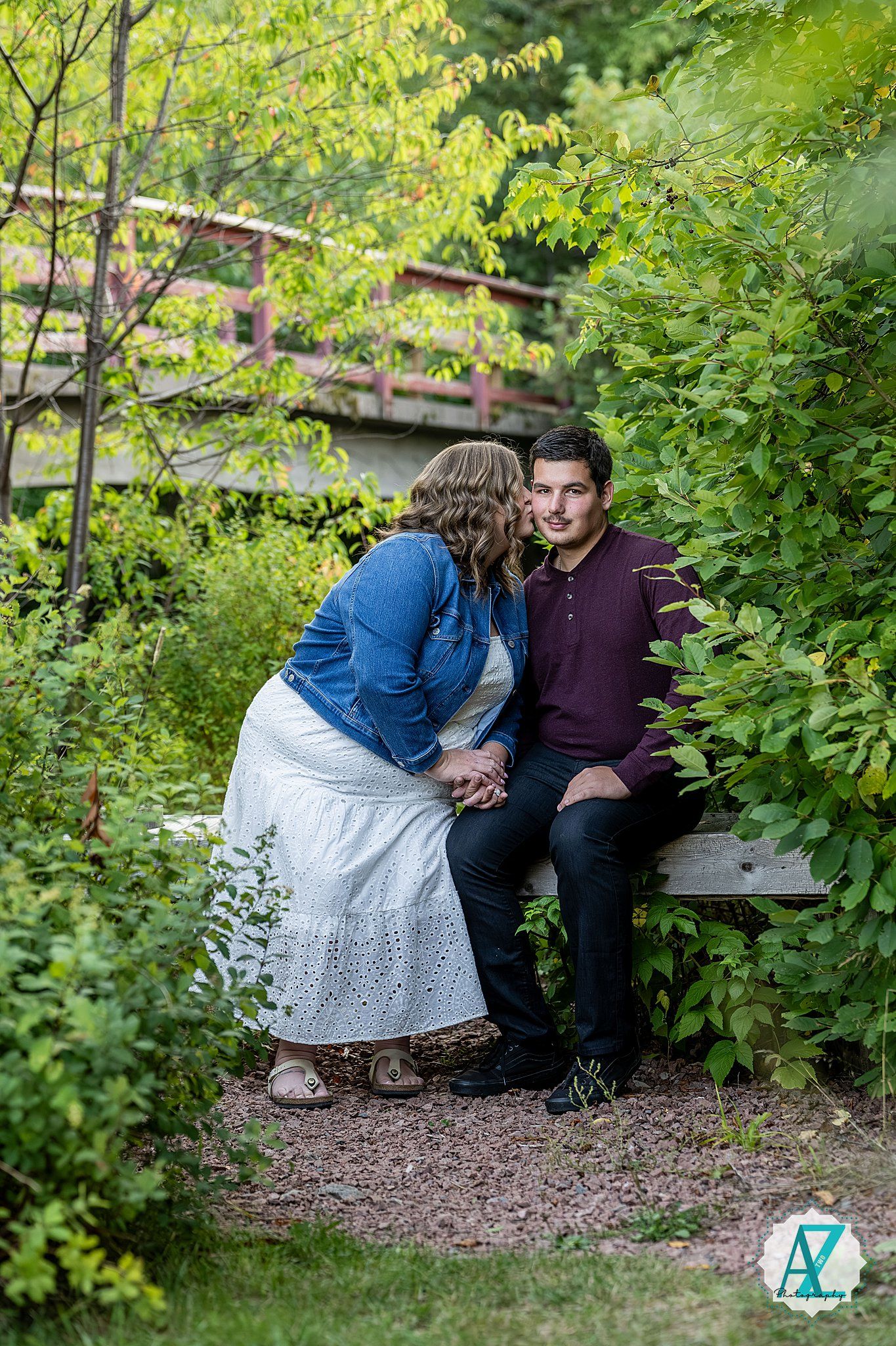 A couple shares an intimate moment while sitting on a bench surrounded by lush greenery in an outdoor setting.