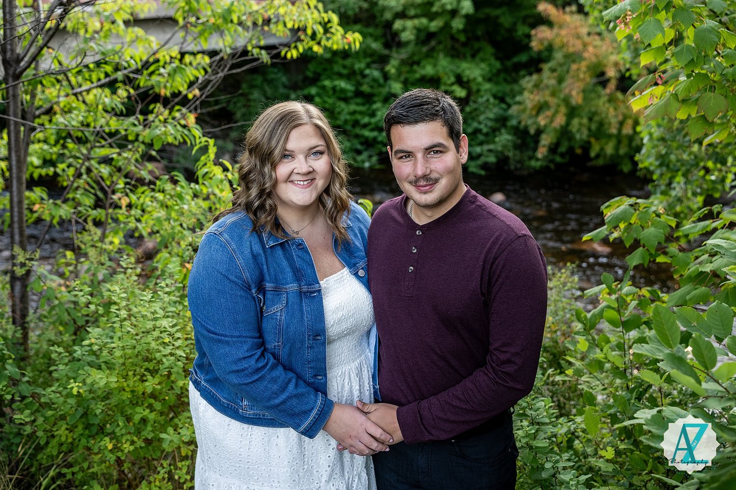 A couple poses together outdoors on a sunny day surrounded by lush green foliage in a garden setting.