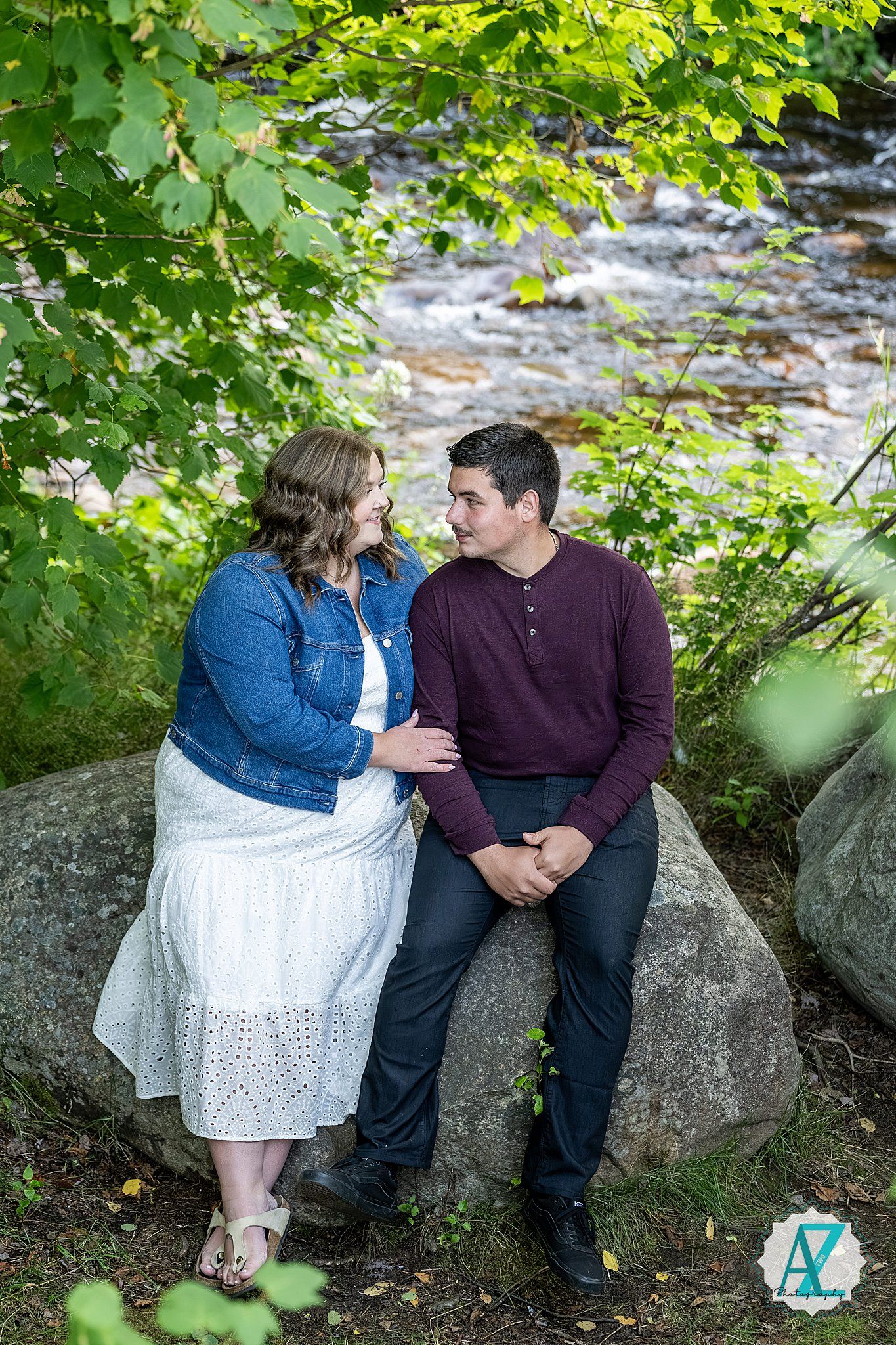 A couple sits together on large rocks by a flowing stream framed by green summer foliage.