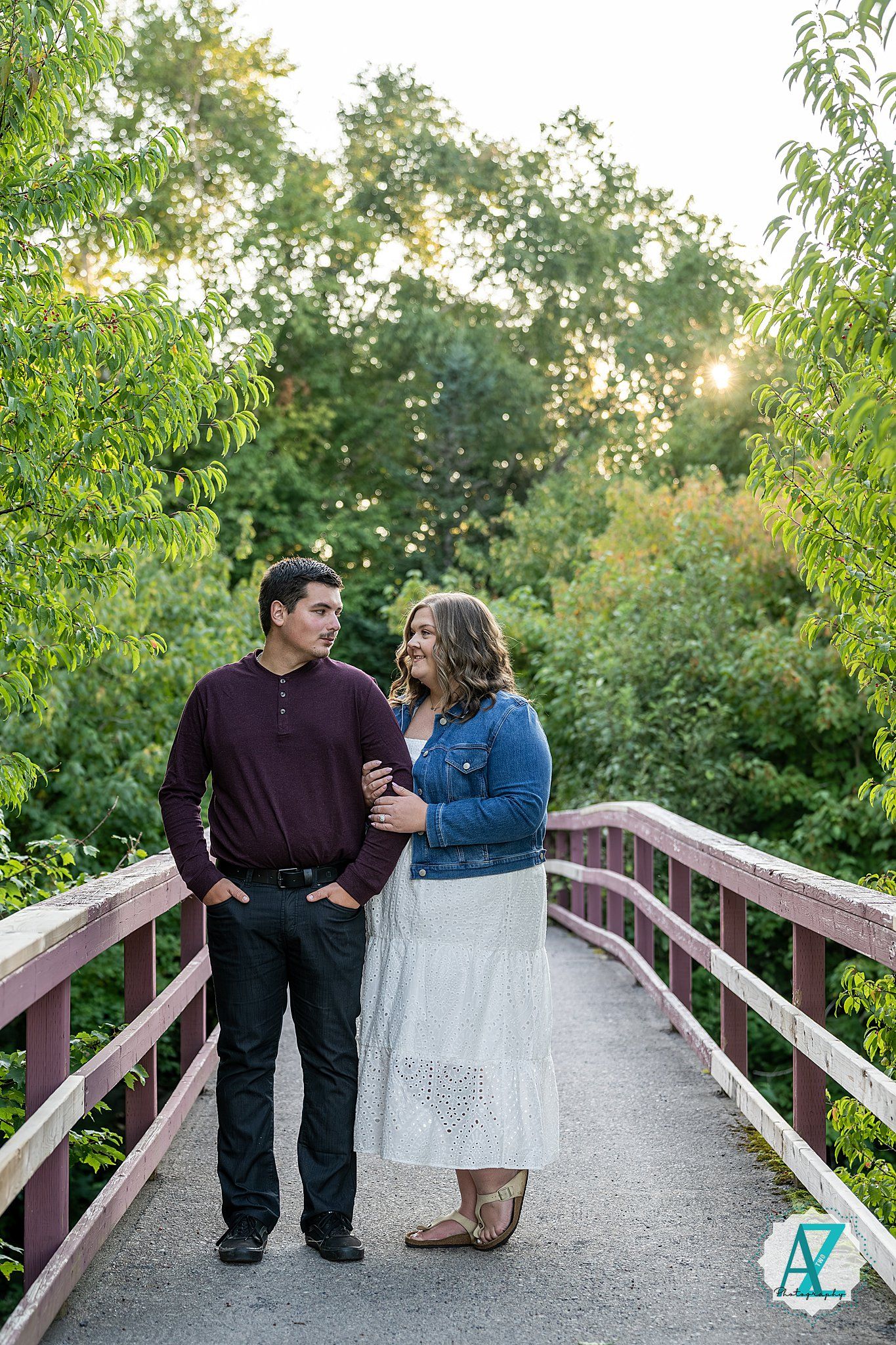 A couple walks together along a wooden footbridge with green trees around them at sunset.