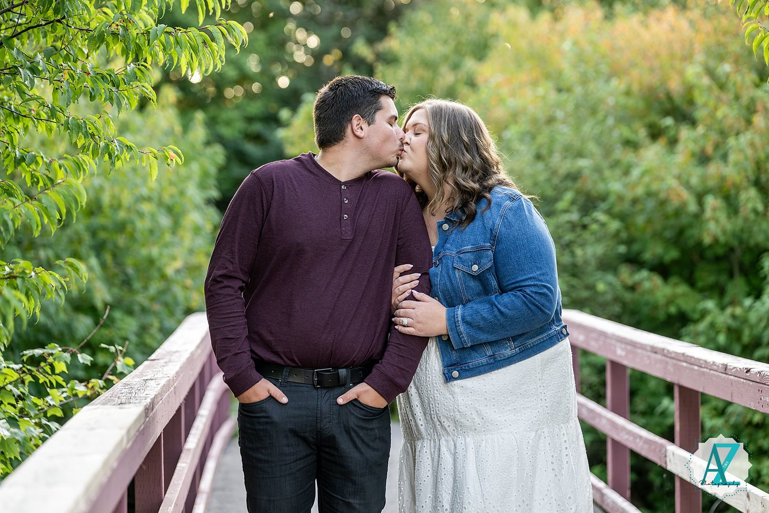 A couple shares a romantic moment on a wooden bridge surrounded by lush summer greenery.
