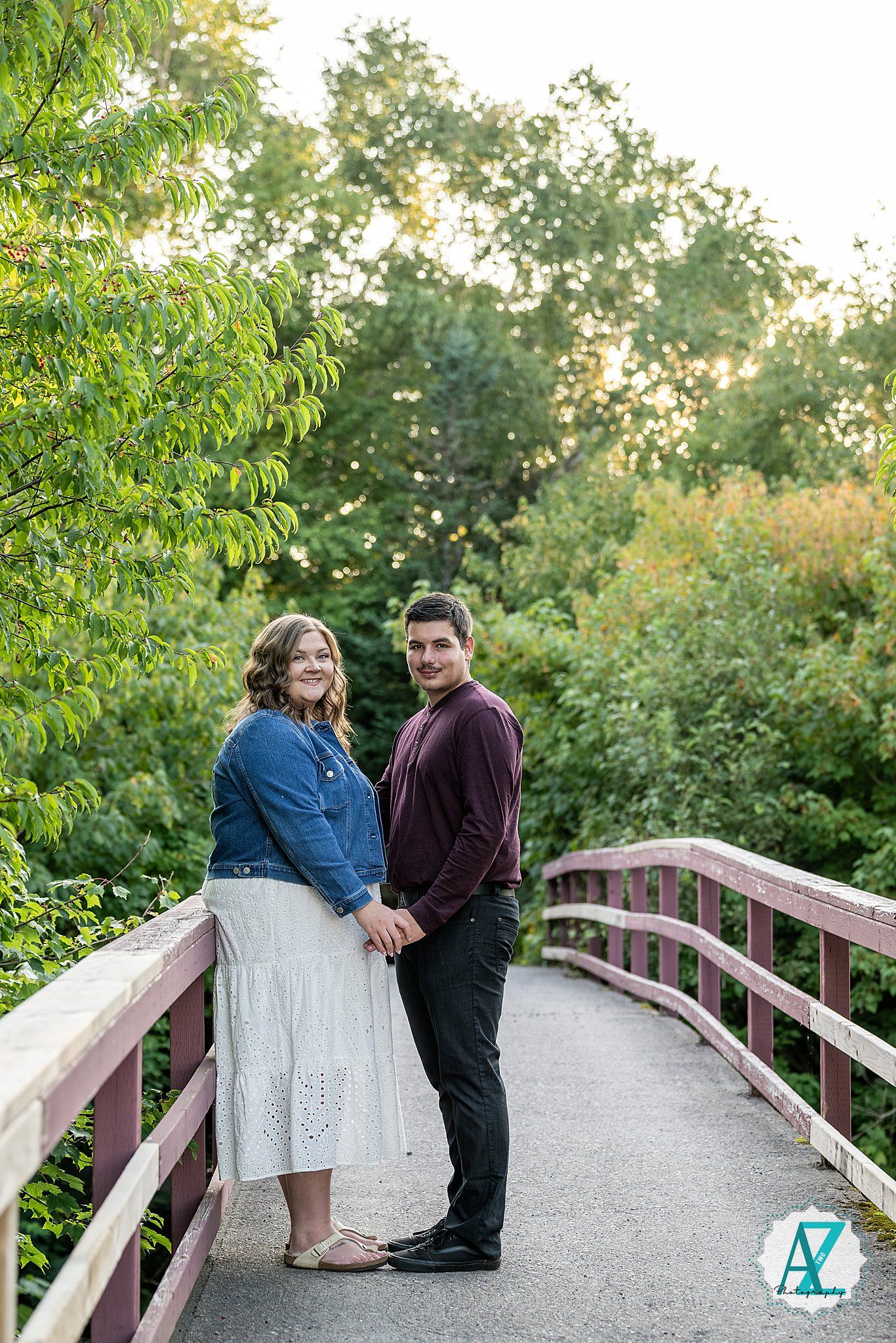A couple shares a quiet moment sitting together on a wooden bridge surrounded by lush green trees.