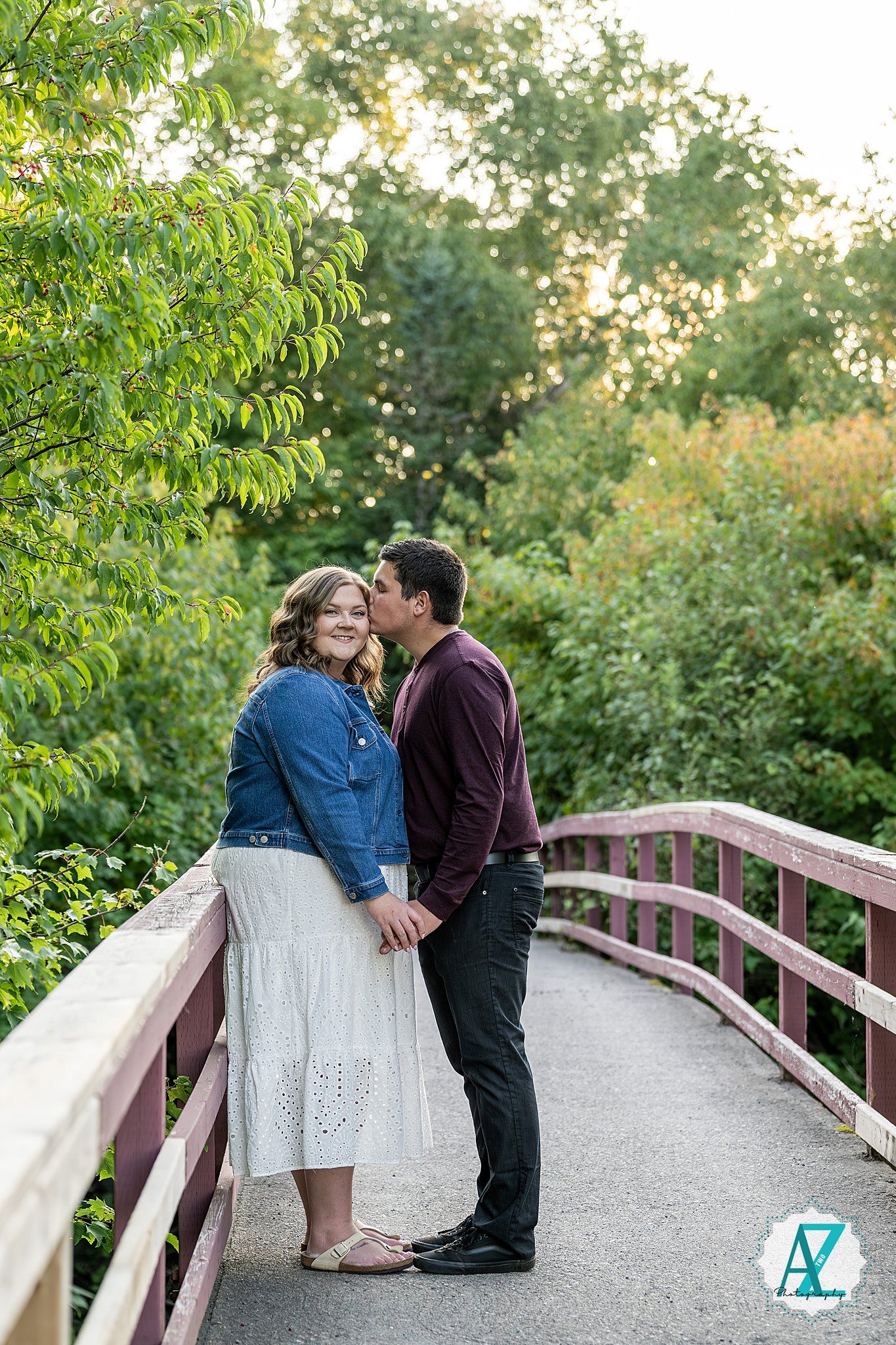 A couple shares a tender moment together on a bridge path surrounded by lush green trees and foliage.