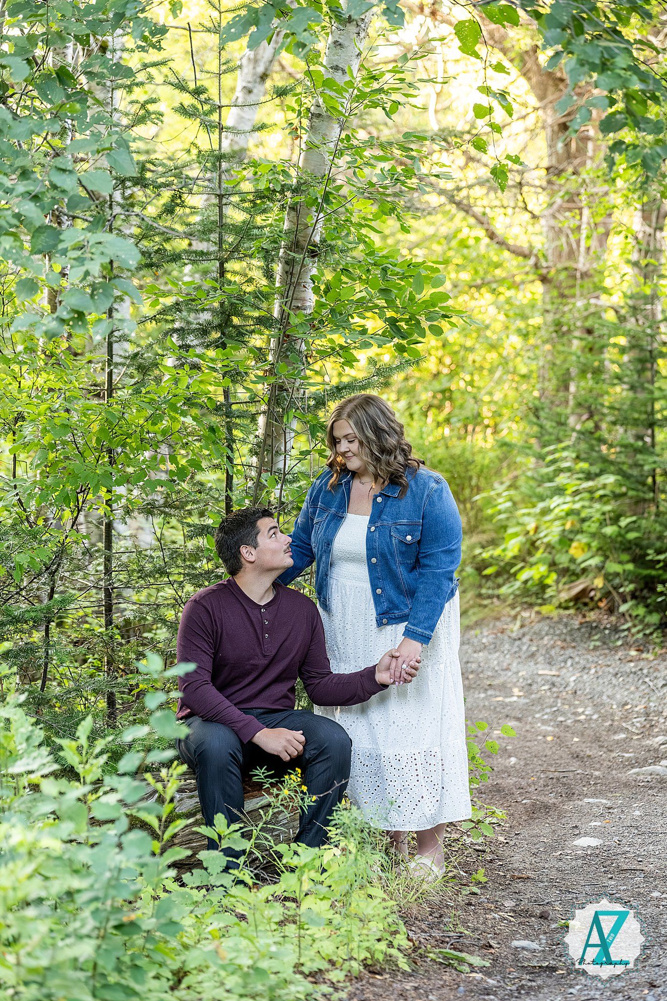 A romantic couple sits together on a forest path surrounded by lush green foliage during a summer photo session.