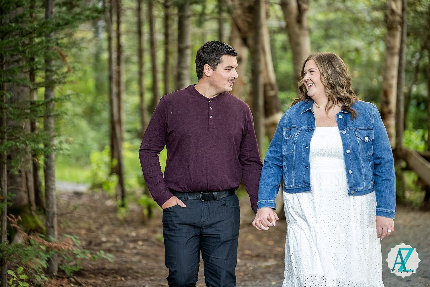 A couple dressed in denim and burgundy enjoys a romantic walk through a lush green forest setting.