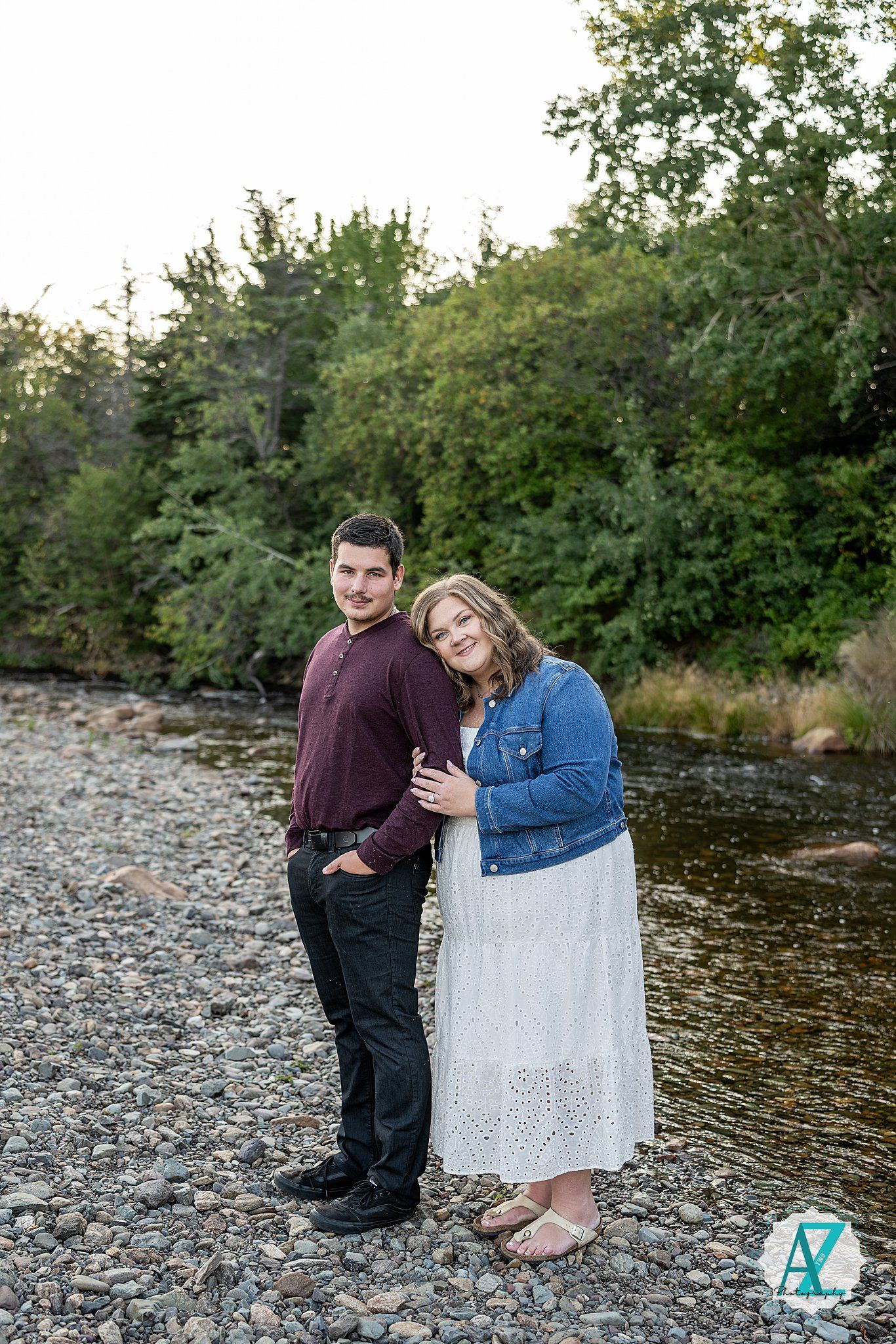 A couple stands together on a rocky riverbank surrounded by greenery during an outdoor photography session.