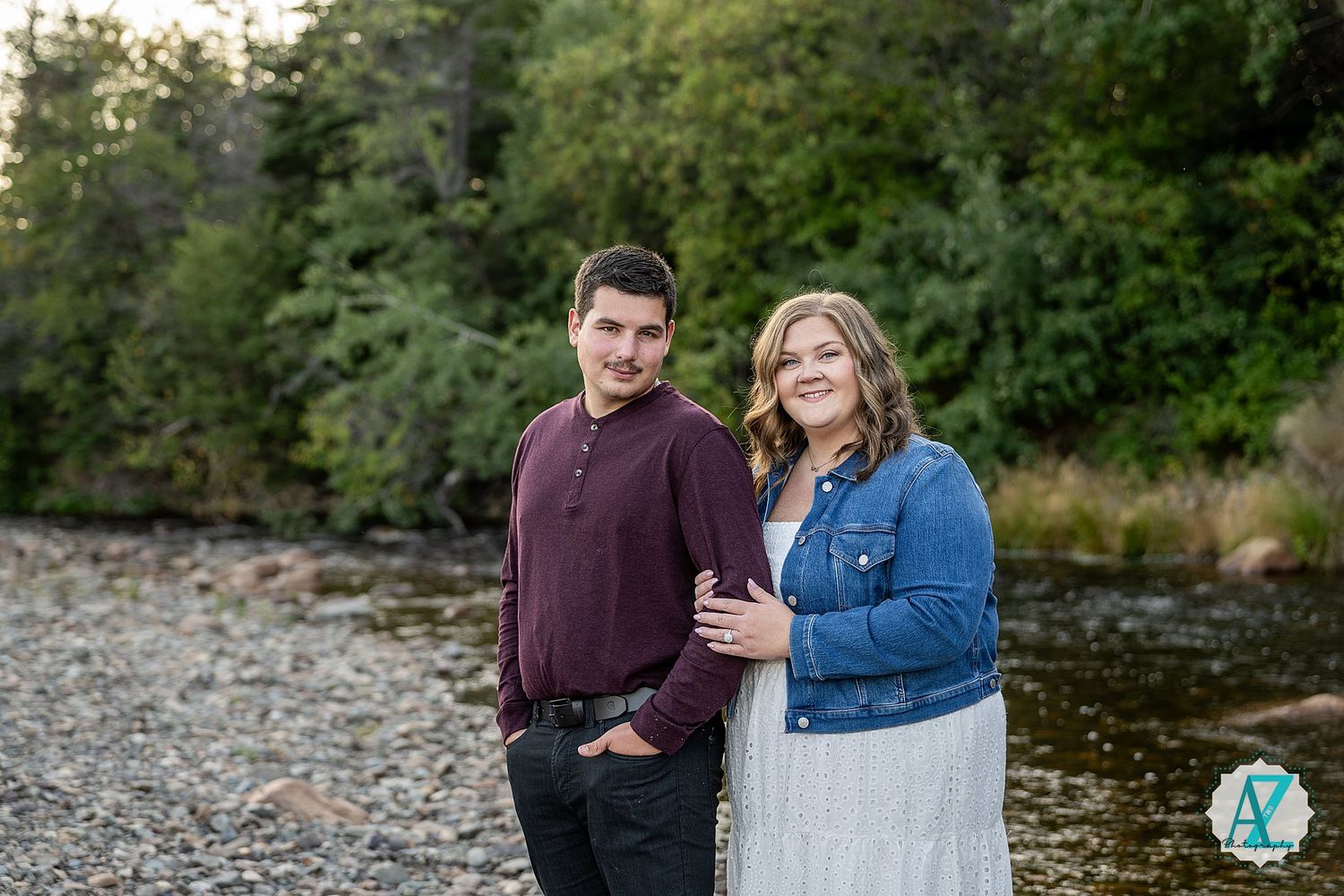 Couple having romantic outdoor photo session near a river during sunset, showcasing natural affection in a wooded setting.