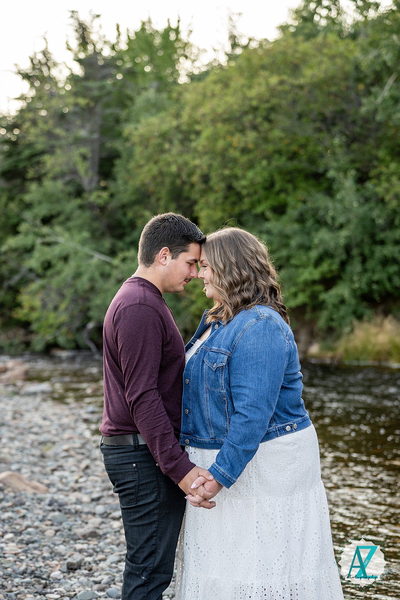 A couple in casual attire shares an intimate moment together by a riverside on a sunny day.