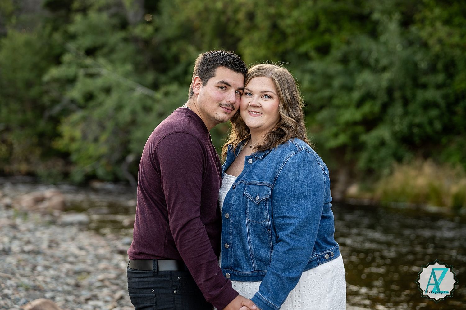 A couple embraces by a river's edge with green foliage in the background during an outdoor photo session.