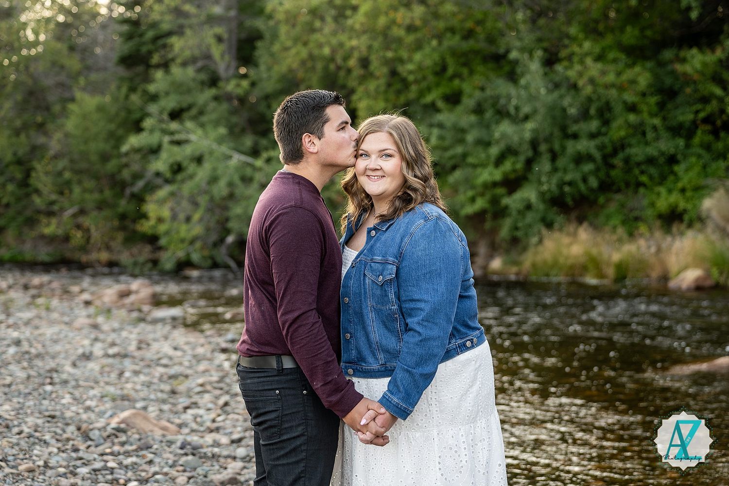 A couple stands together holding hands by a riverbank on a sunny autumn day wearing maroon and denim.