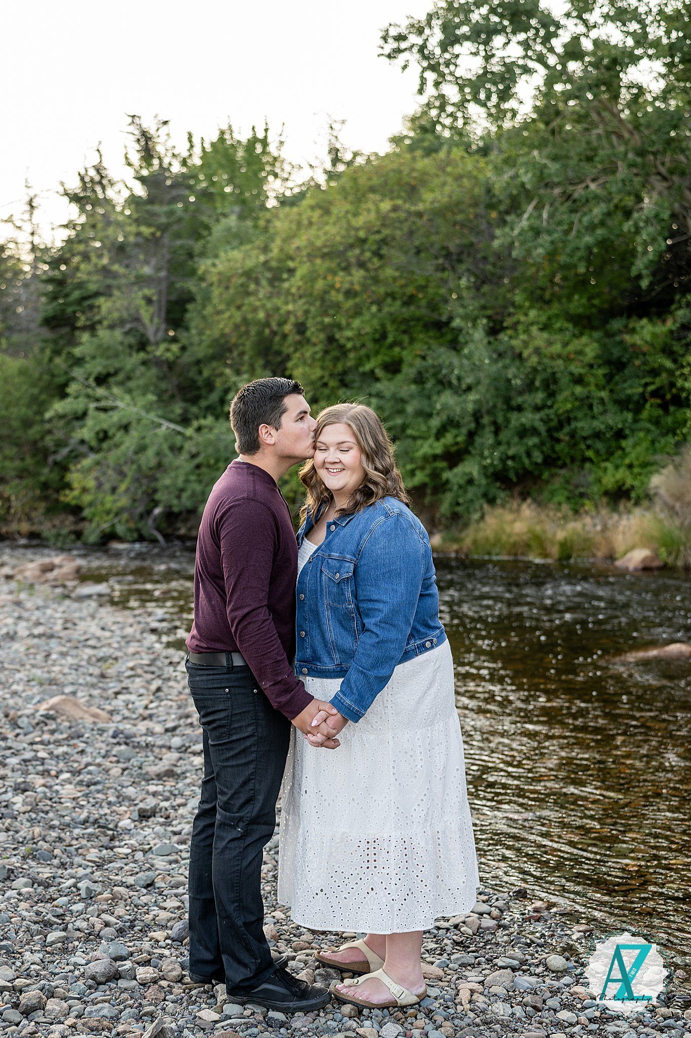 Romantic couple shares tender moments by a riverbank surrounded by lush greenery during outdoor photo session.