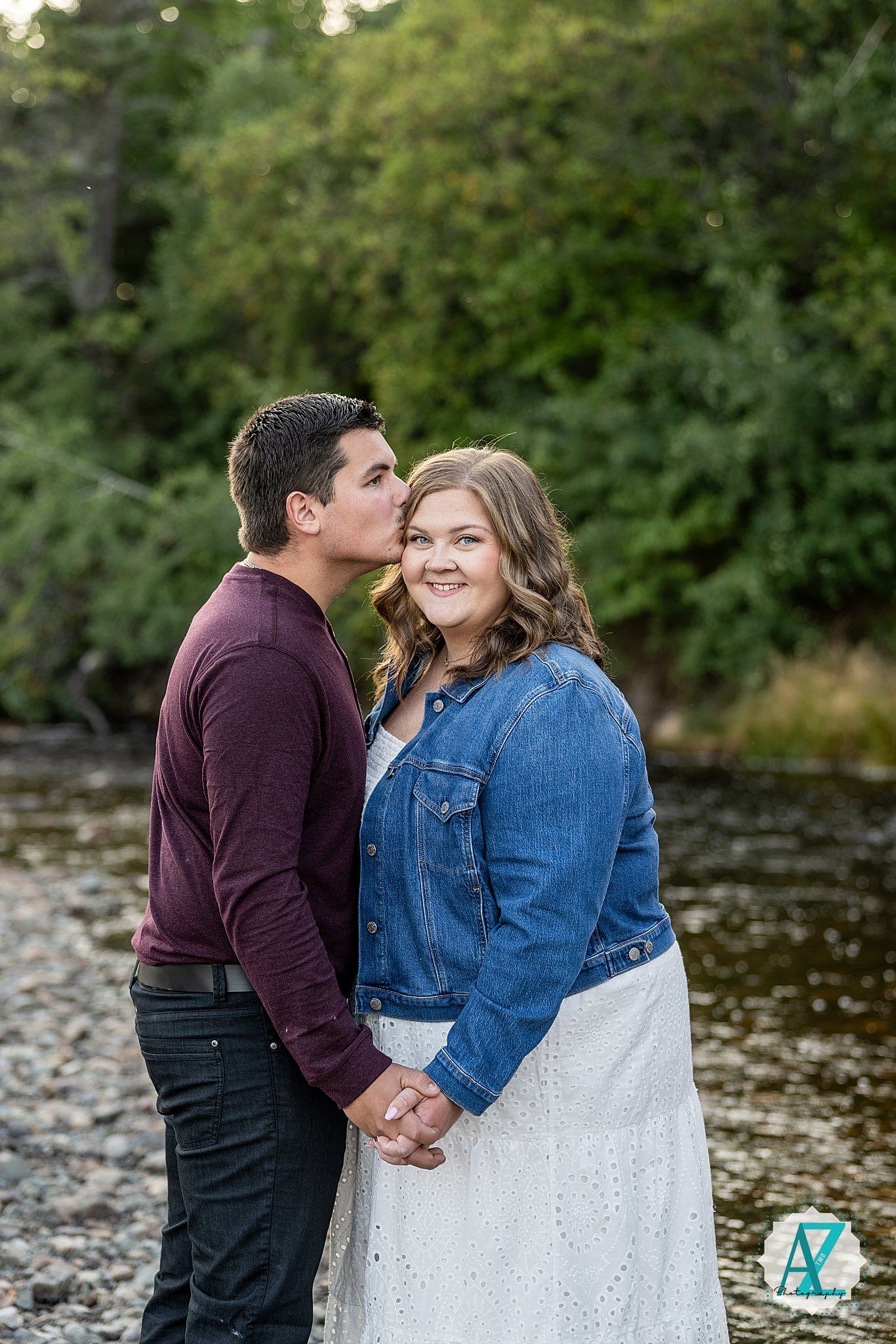A couple sits by a water's edge in autumn, dressed in burgundy sweater and denim jacket.