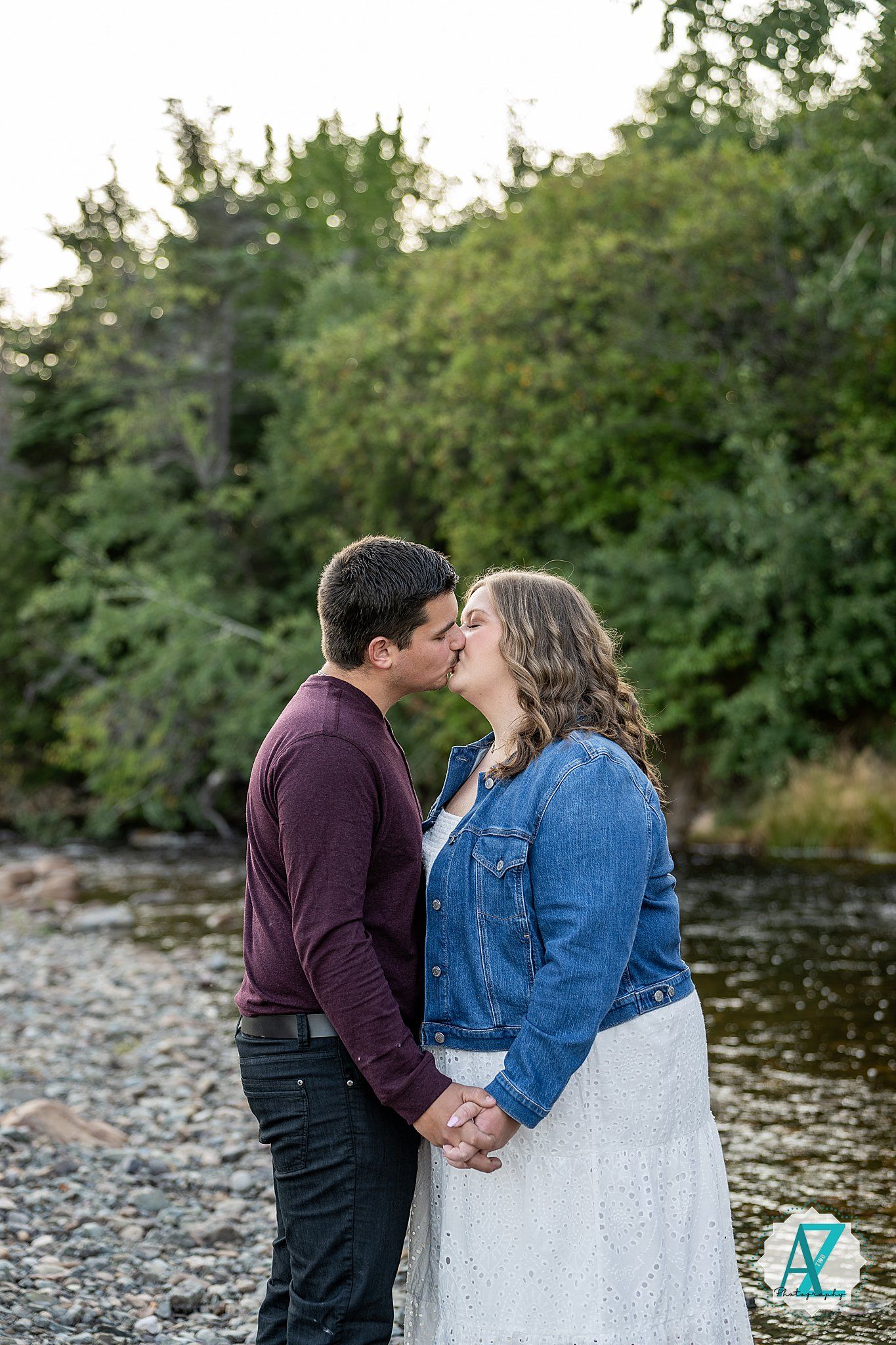 A couple shares a romantic kiss by a riverside with lush greenery in the background during golden hour.