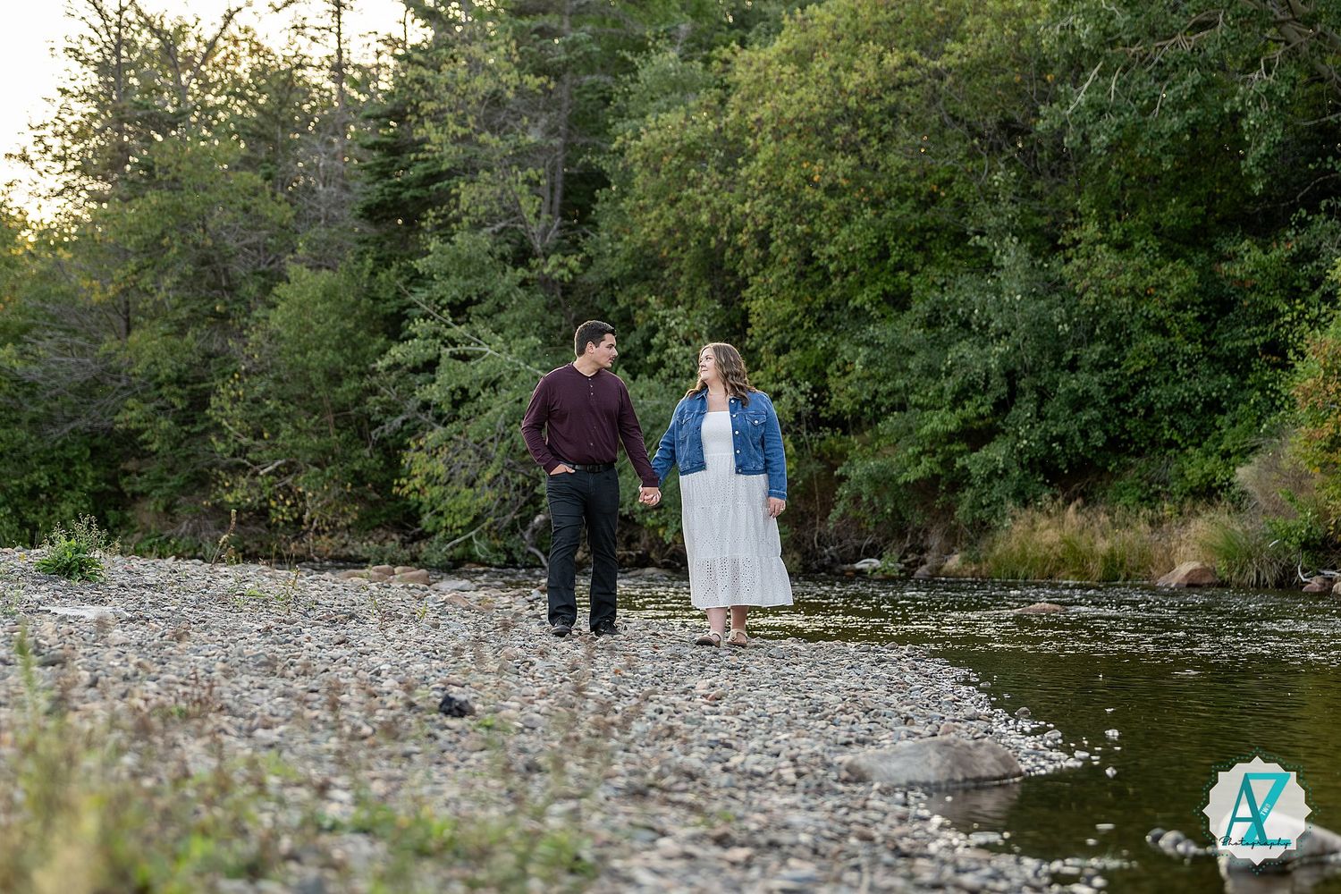 Two people walk along a rocky riverbank surrounded by lush green trees on a sunny day.