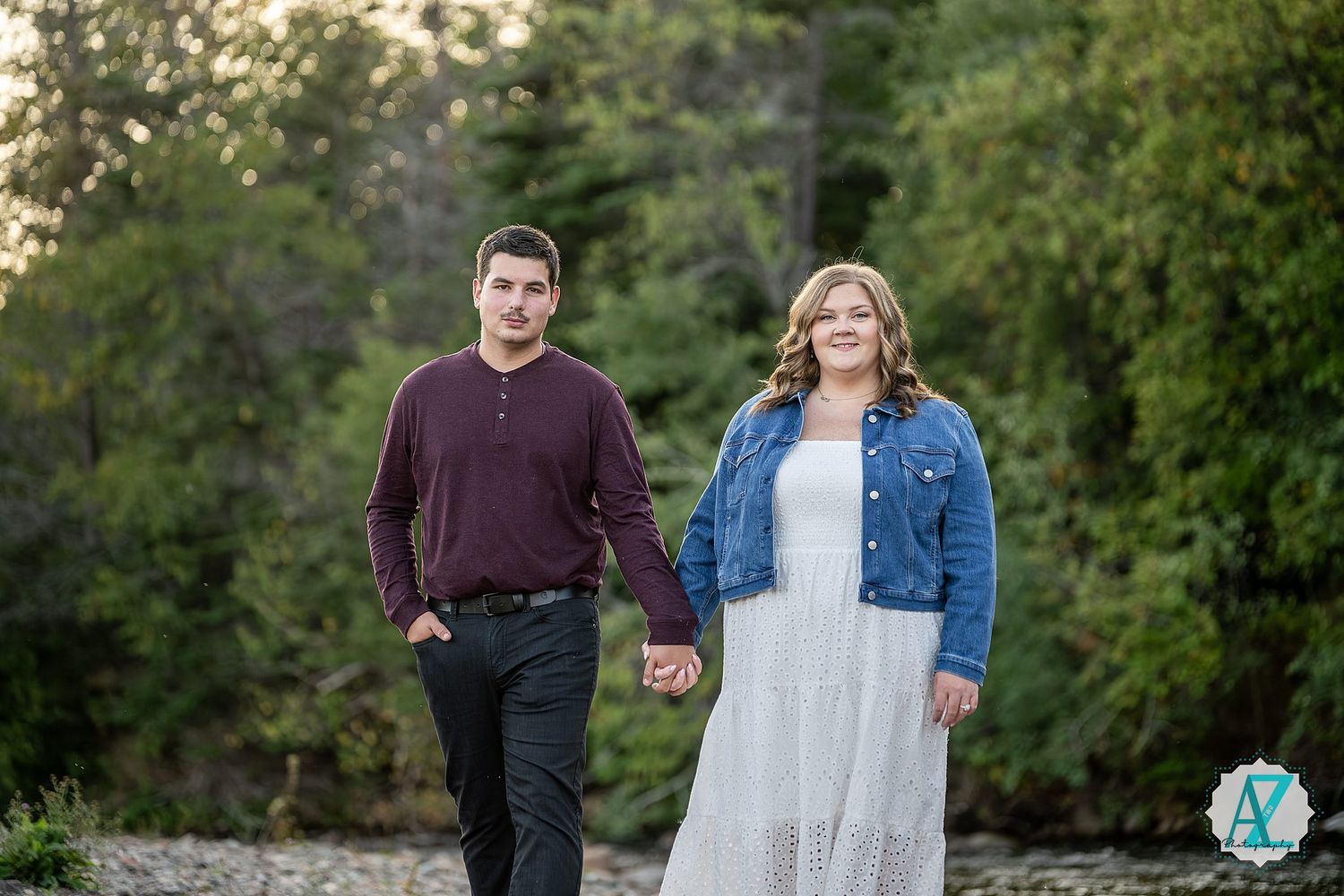 A couple holding hands walks together outdoors, with her wearing a denim jacket over white dress and him in a burgundy shirt.